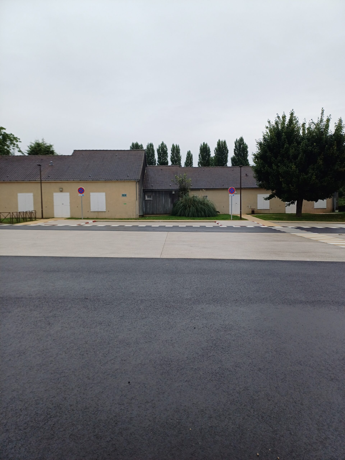 Bâtiment beige avec portes blanches, parking et arbres.