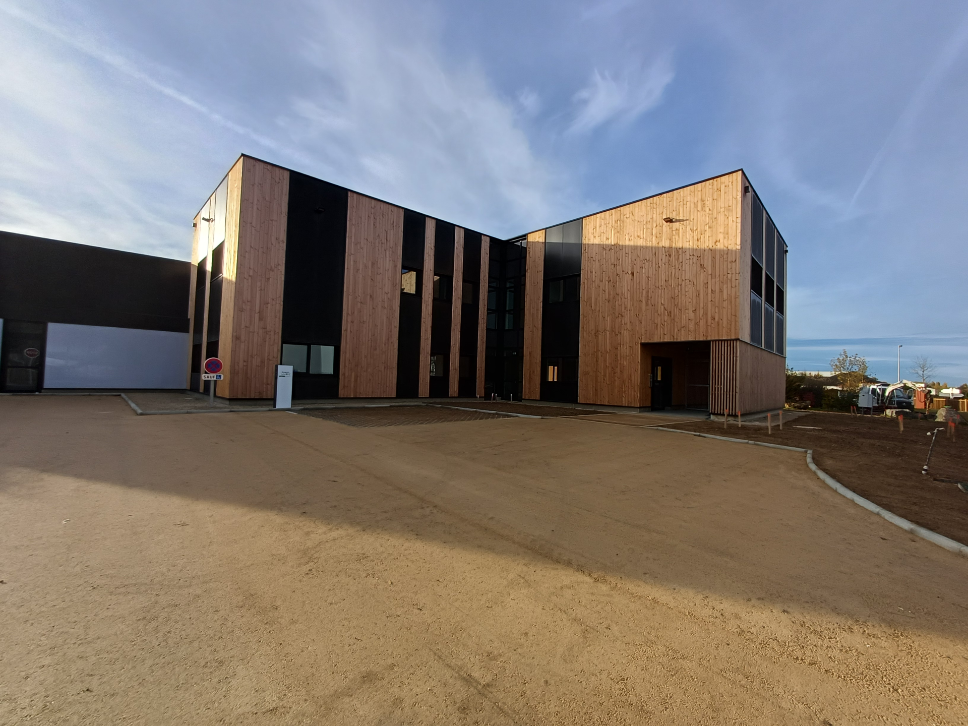 Bâtiment en bois avec grandes fenêtres, parking sableux, ciel bleu.