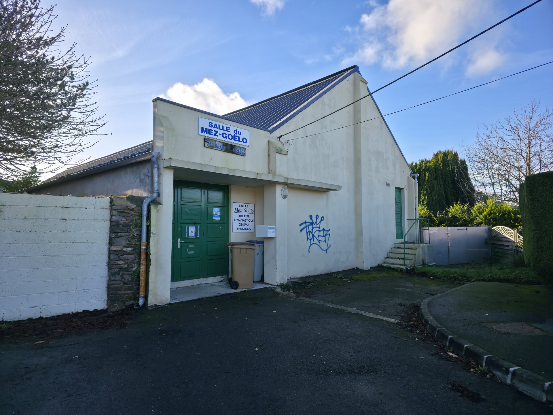 Un petit bâtiment avec un panneau indiquant "Salle de Mez-Kelo". Porte verte, poubelles, graffiti, mur de pierre, allée.