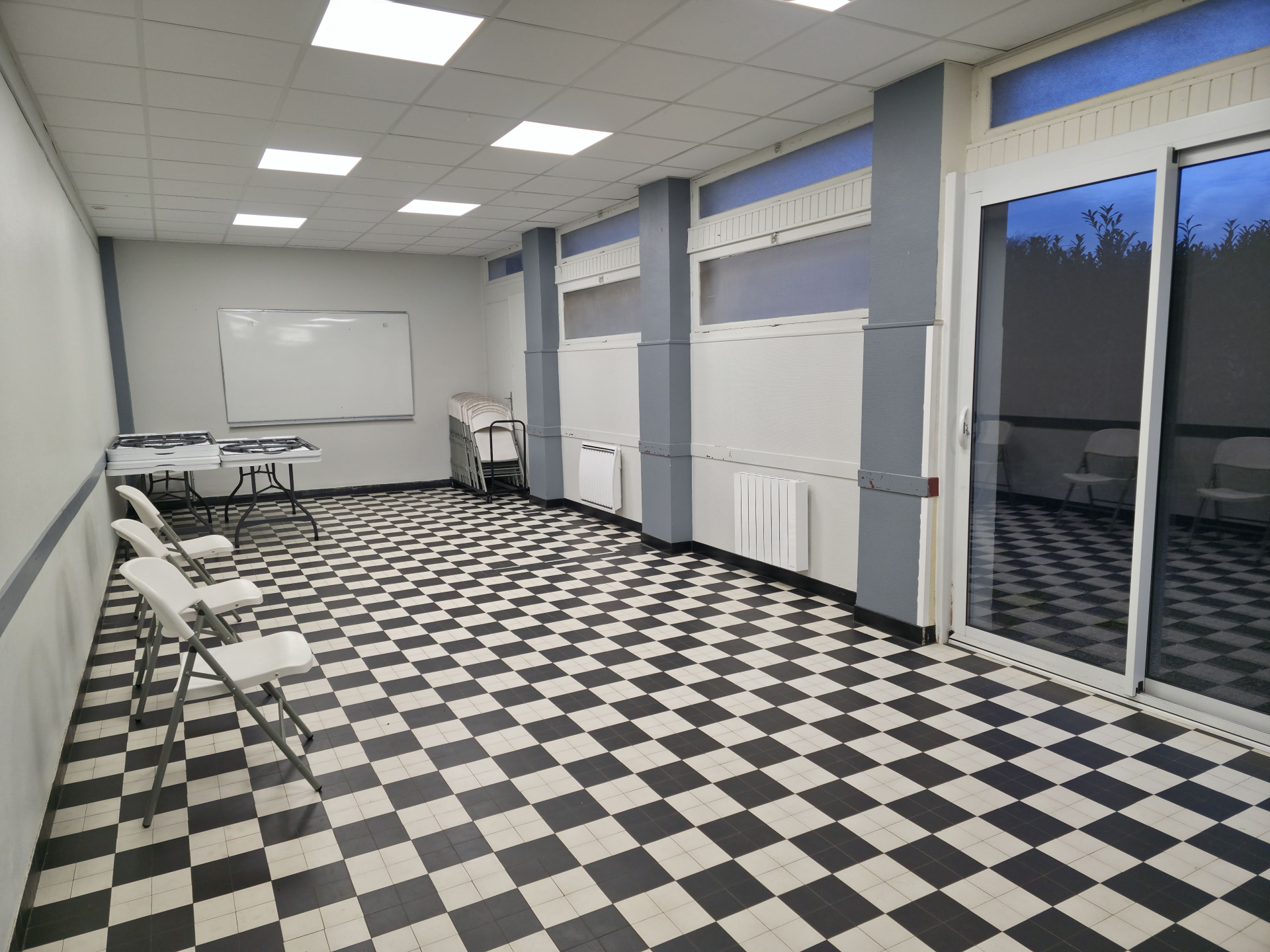 Salle avec table, chaises, tableau blanc, radiateurs, porte vitrée et sol en damier noir et blanc.