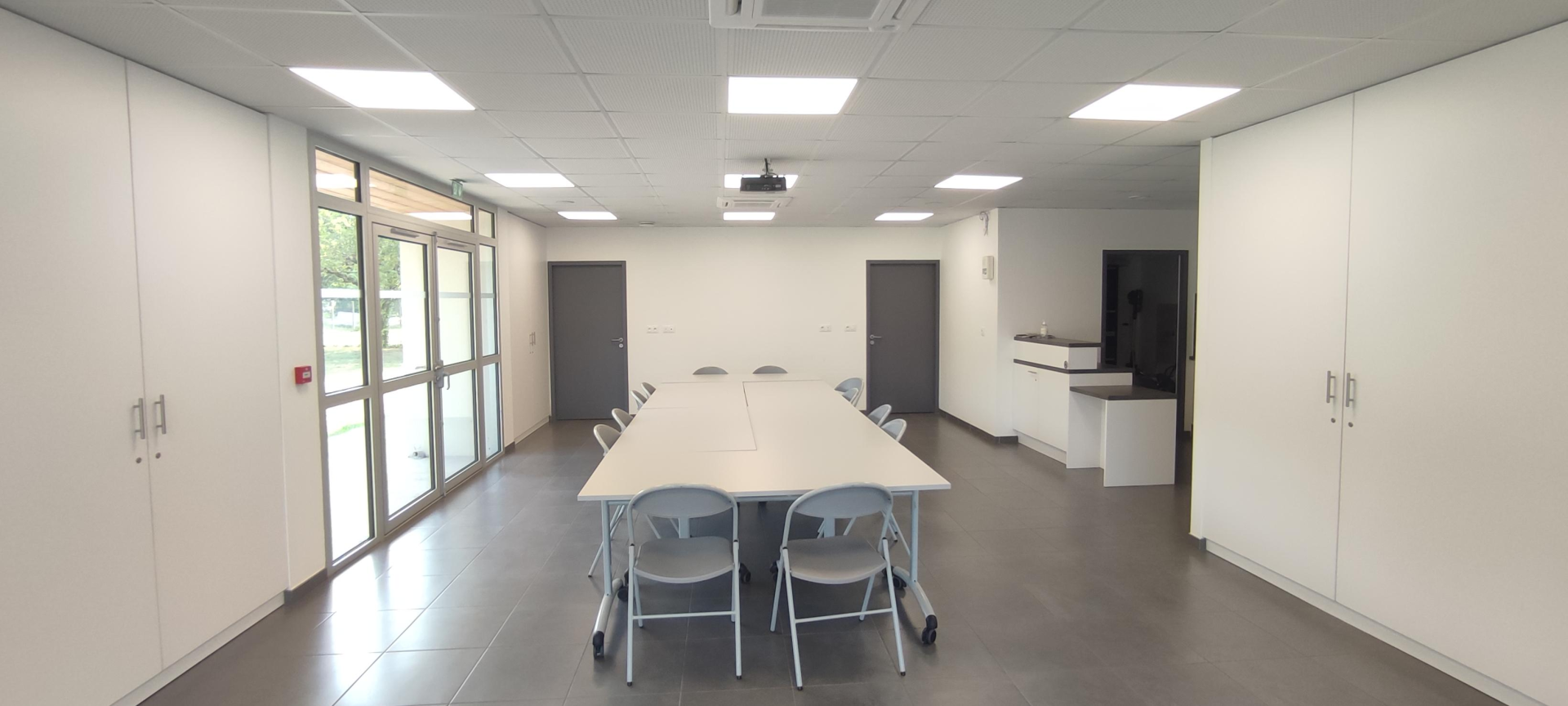 Salle de réunion avec table, chaises, vidéo projecteur, placards, portes et fenêtres.