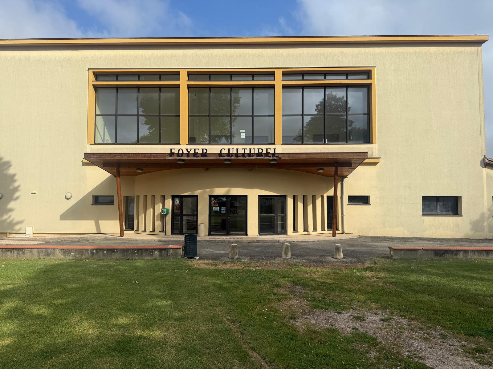 Bâtiment avec entrée, grandes fenêtres, enseigne "Foyer Culturel", pelouse et ciel.