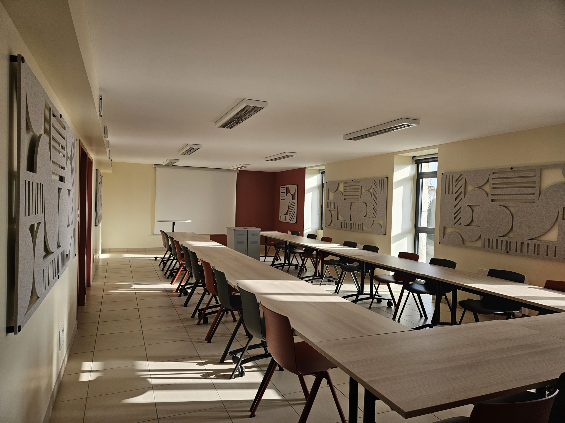 Salle de classe avec deux longues tables, chaises, tableaux et lumière naturelle.