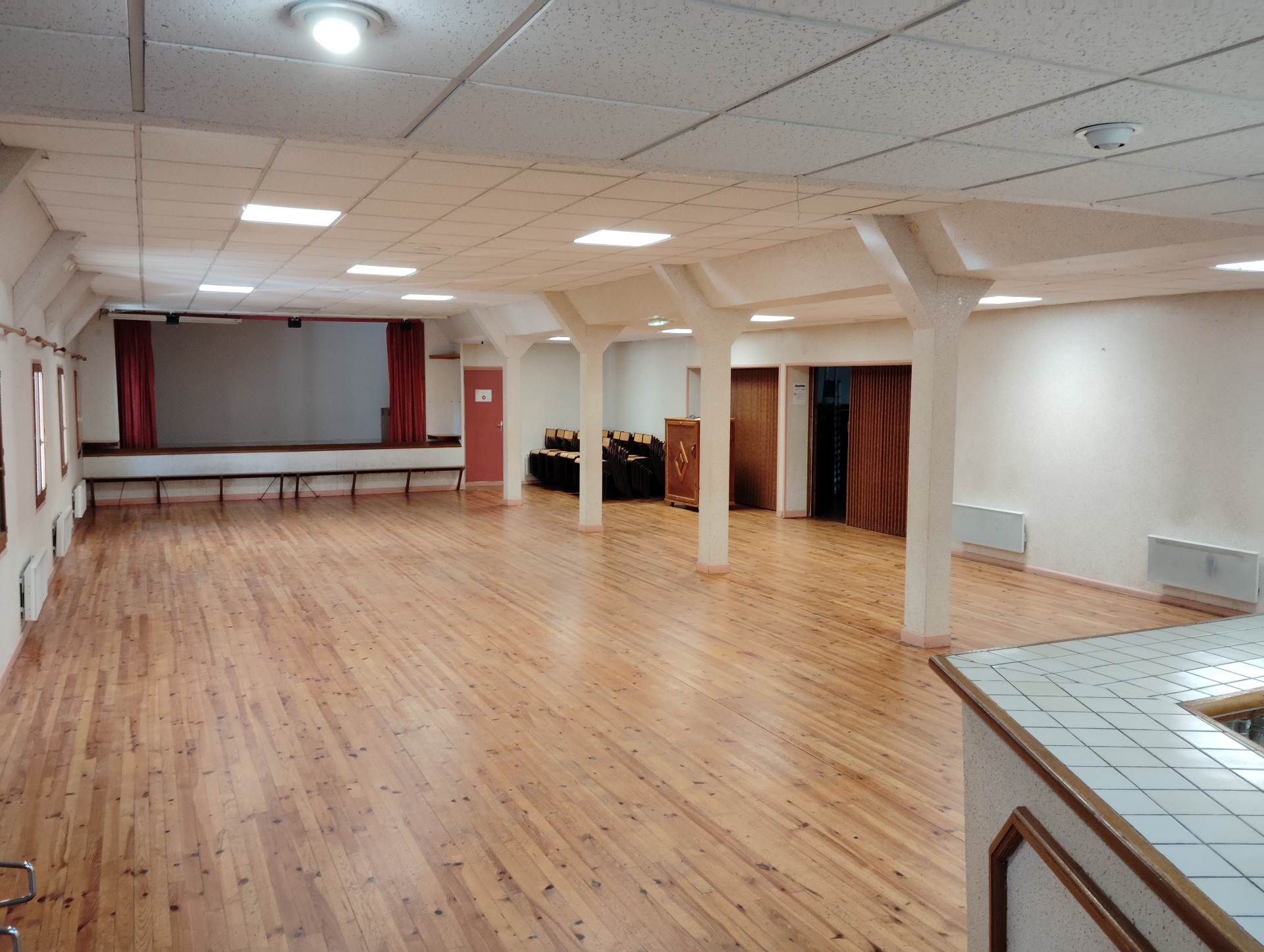 Salle spacieuse avec parquet, tables, chaises, rideaux, et portes.