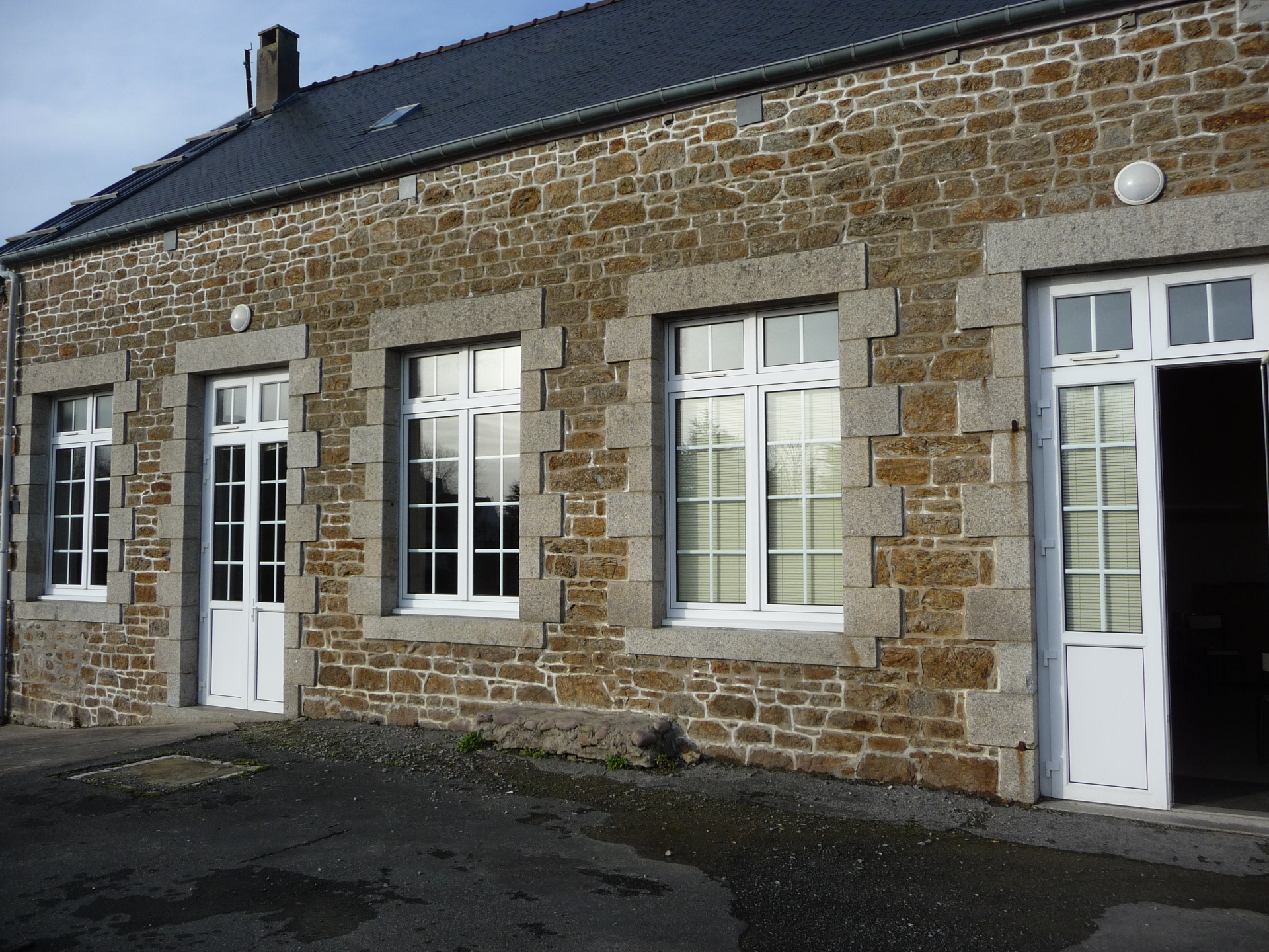 Maison en pierre avec portes et fenêtres blanches, et toit à tuiles sombres.