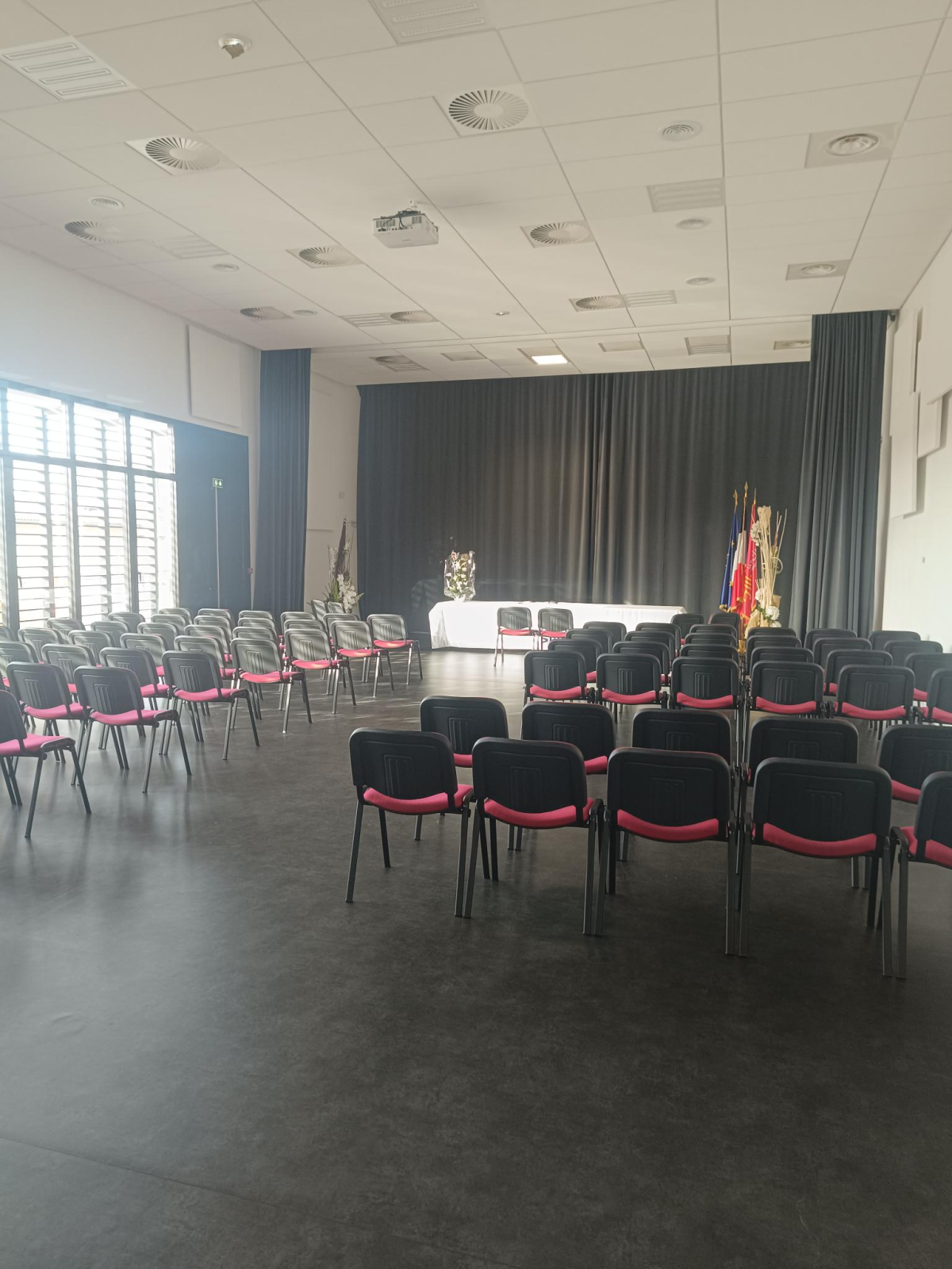 Salle avec chaises alignées, podium, drapeaux, fenêtres et stores.