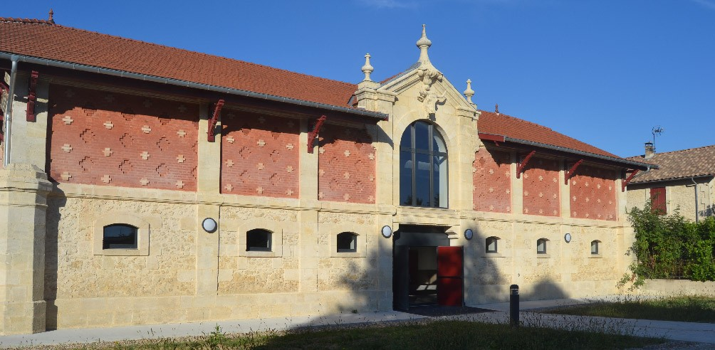 Bâtiment avec toiture en tuiles rouges, murs en pierre et briques, porte centrale rouge et fenêtres de chaque côté.