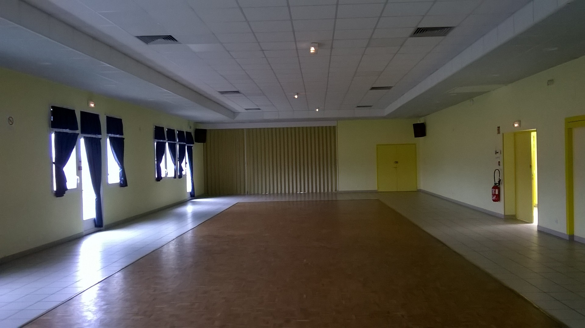 Salle vide avec fenêtres, porte, extincteur, et projecteur plafond.
