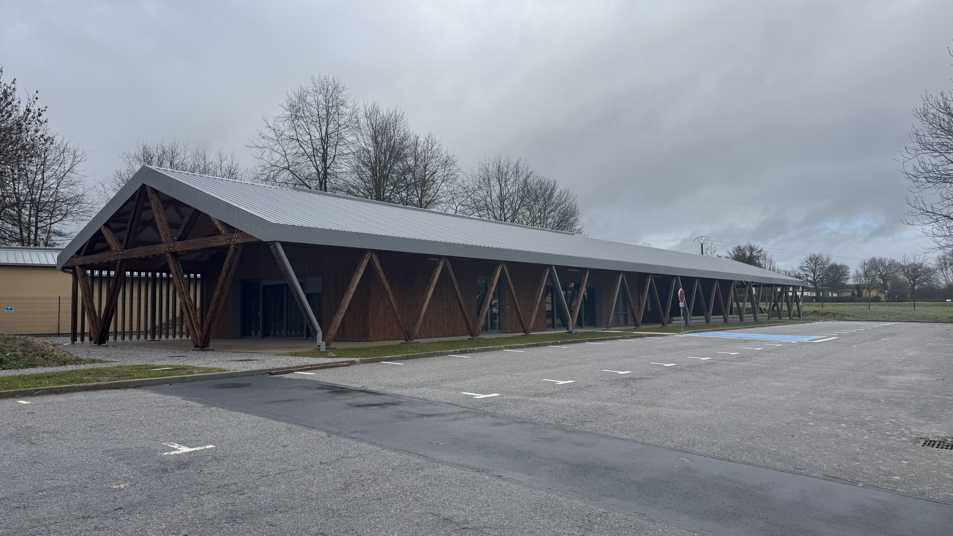 Bâtiment long avec toit incliné, structure en bois et grandes fenêtres, stationnement devant.