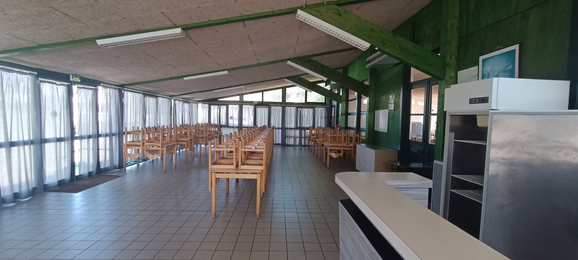 Cafétéria avec tables, chaises, frigo, comptoir, et fenêtres.