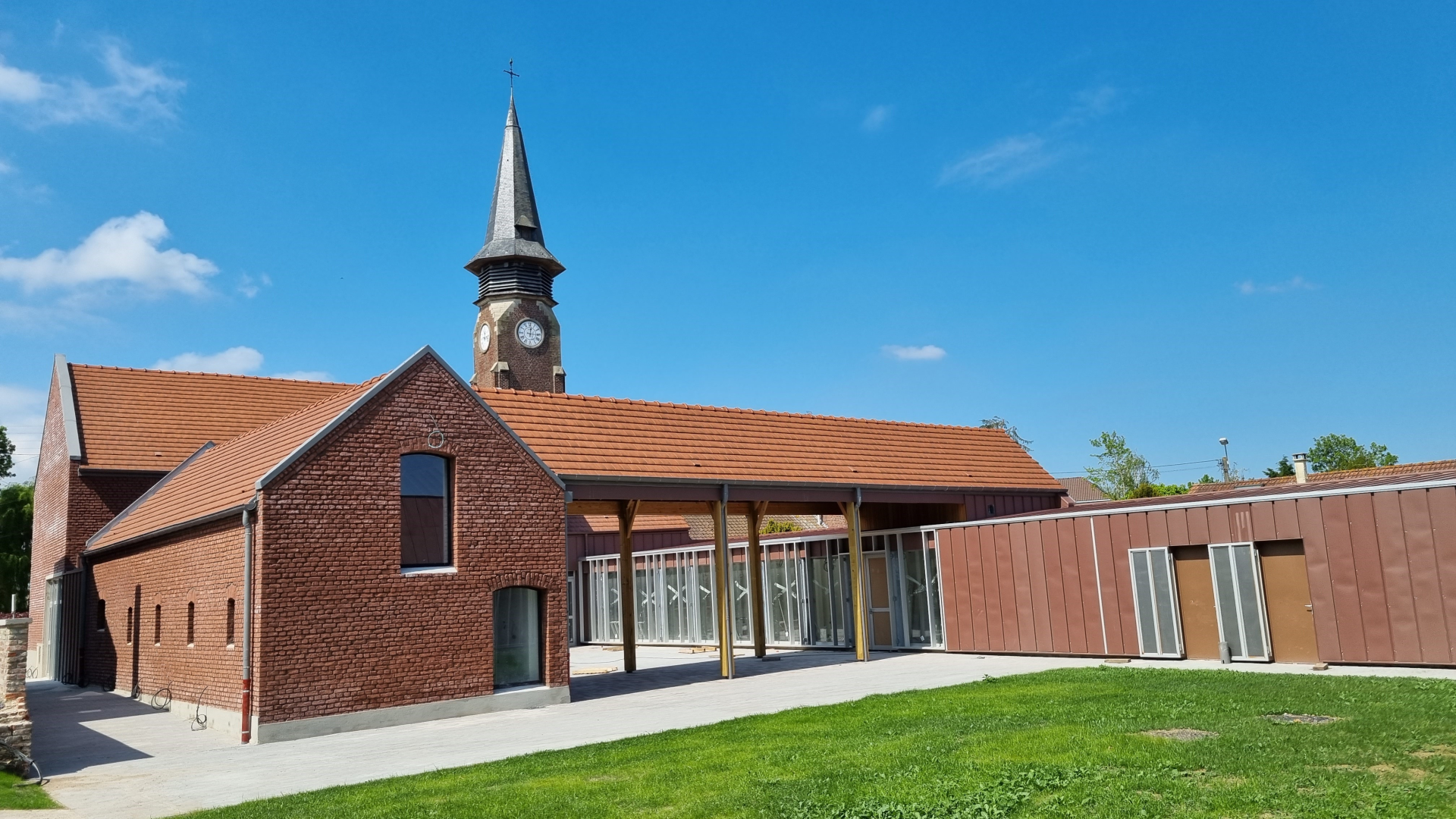 Église avec clocher, toit en tuiles, murs en briques, entrée vitrée et pelouse.