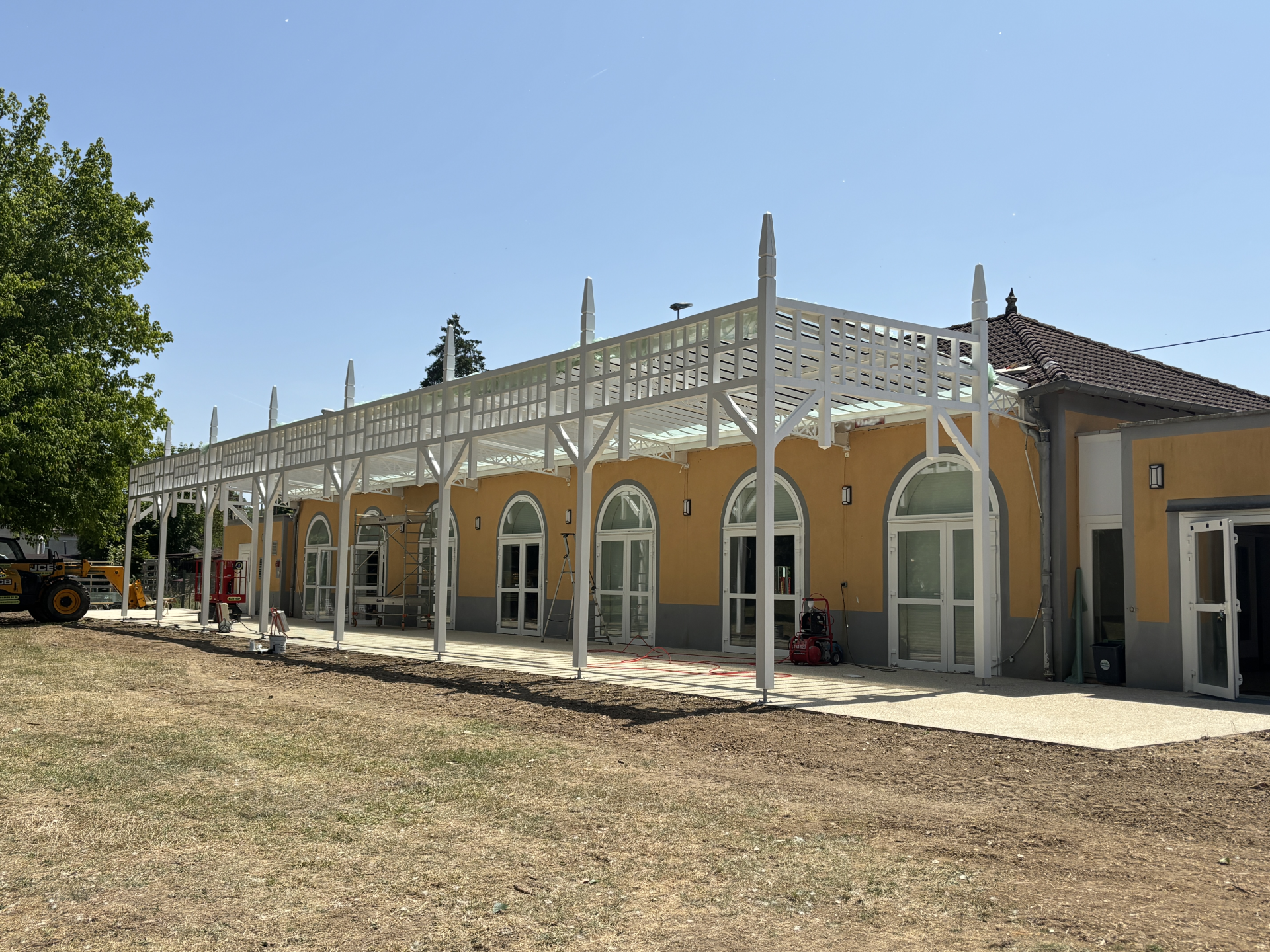 Bâtiment avec structure blanche et murs jaunes, portes vitrées, toit en tuiles, et terrain herbeux.