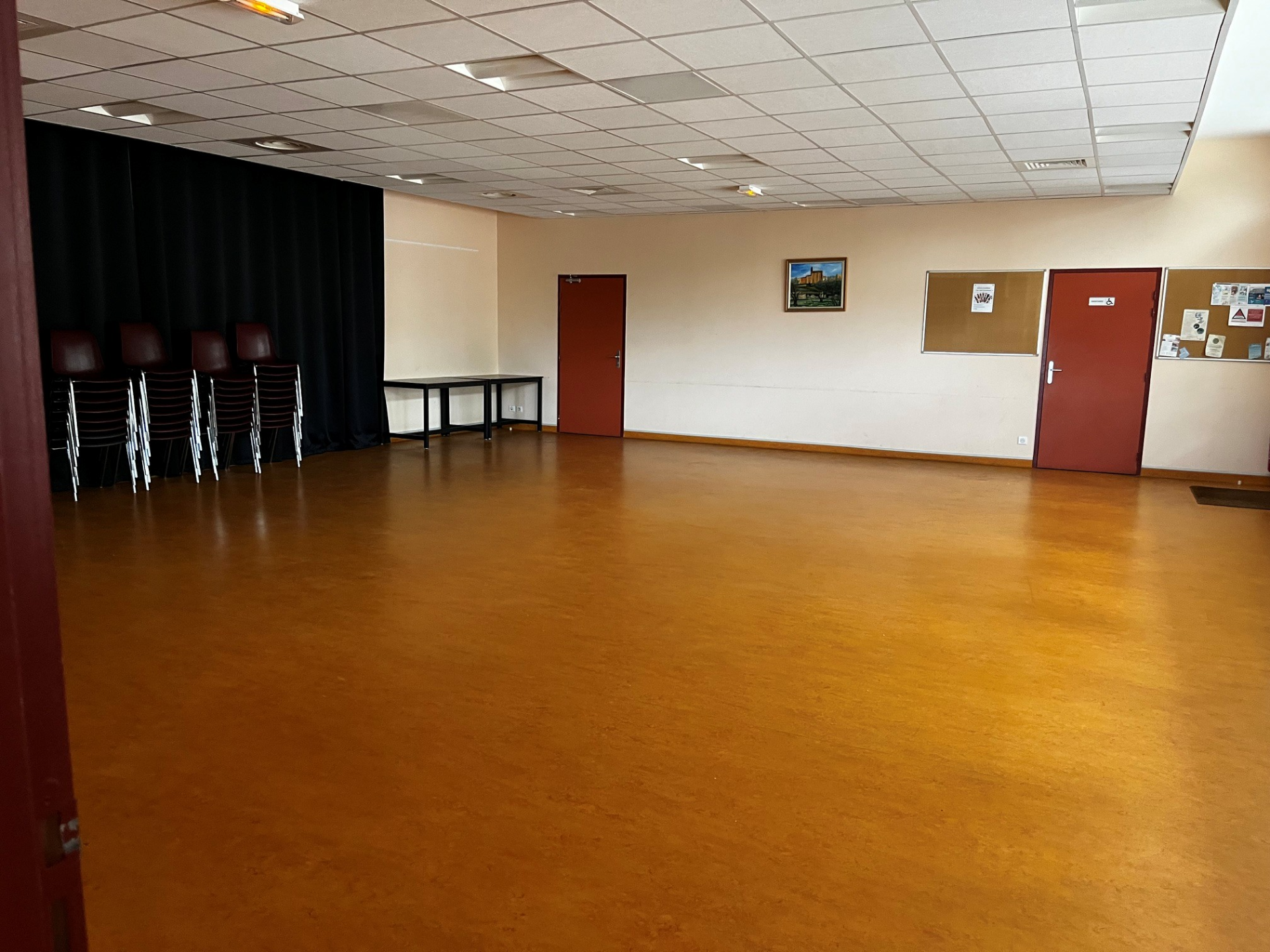 Salle avec parquet, chaises empilées, table, porte rouge, tableau d'affichage et rideau noir.