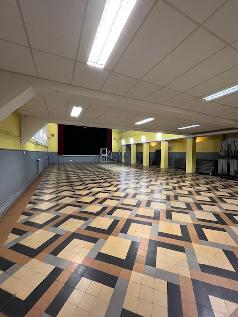 Grande salle avec motif de sol carrelé, éclairage au plafond, murs jaunes et porte au fond.