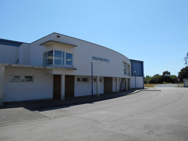 Bâtiment avec enseigne "Salle des Fêtes", parking vide, ciel dégagé.