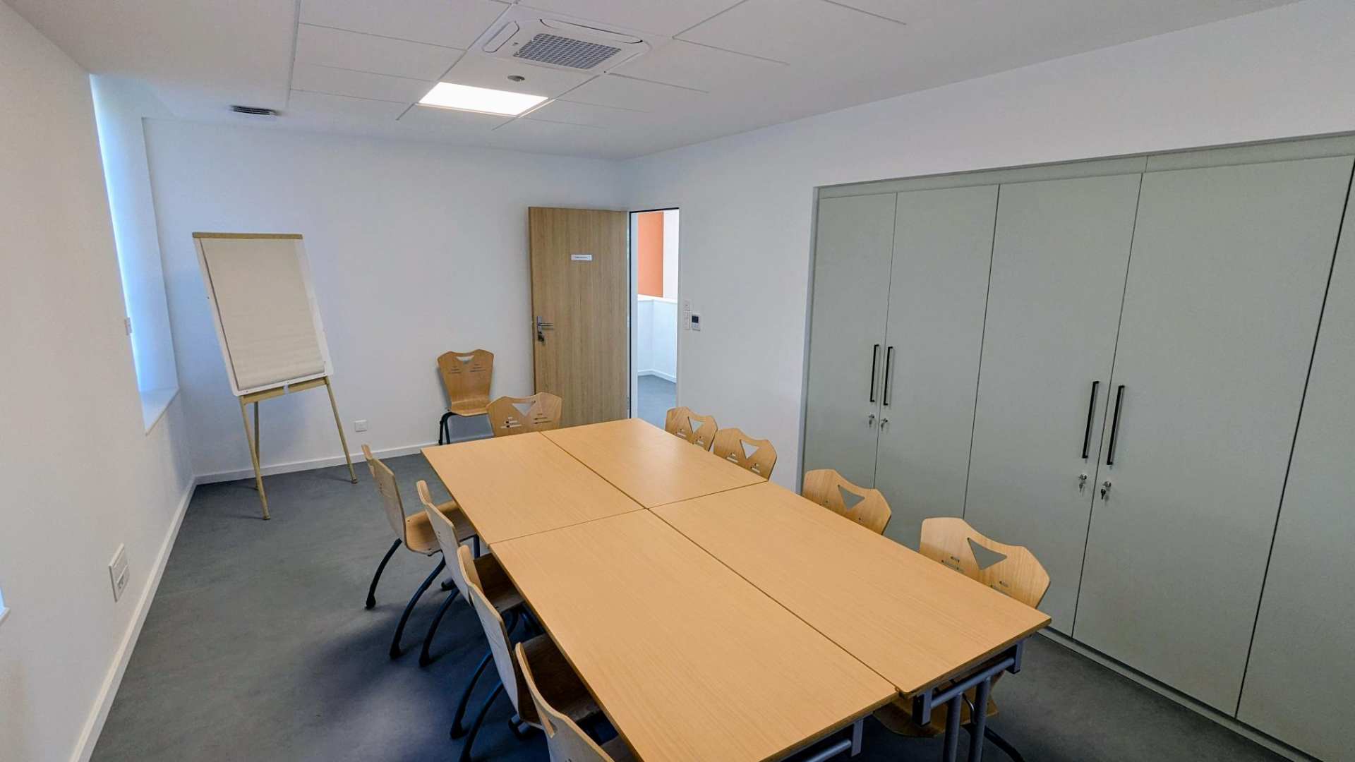 Salle de réunion avec table, chaises, tableau blanc, armoire et porte.