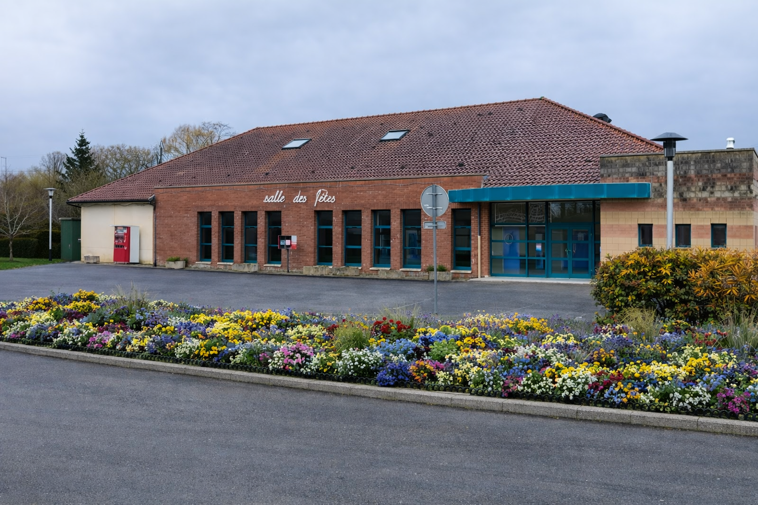 Bâtiment avec toiture en tuiles, murs en brique, grandes fenêtres, entrée avec auvent bleu, panneau de signalisation, parterre de fleurs.