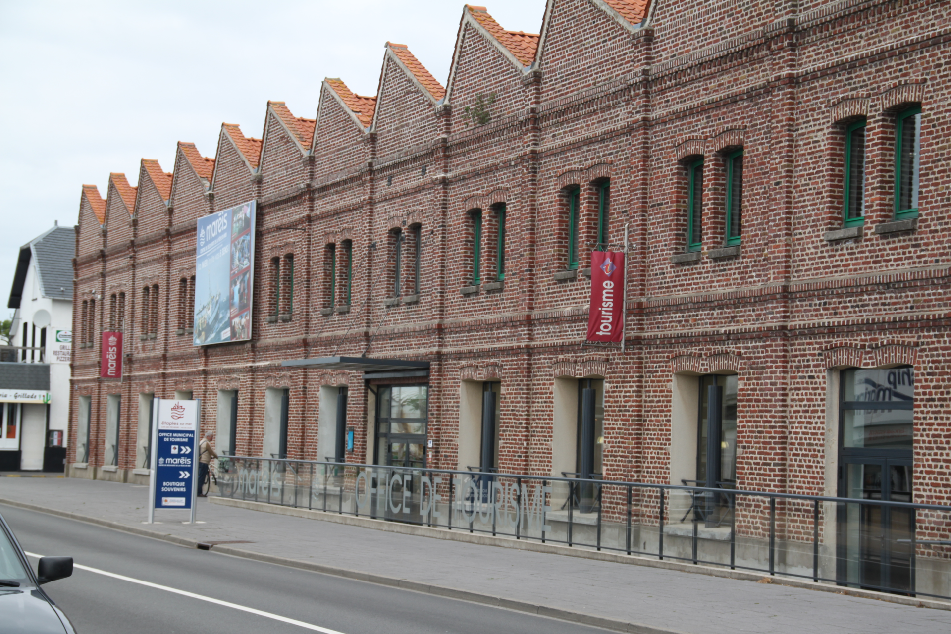 Bâtiment en briques avec toits en croupe, fenêtres vertes, bannières publicitaires et barrières le long de la route.