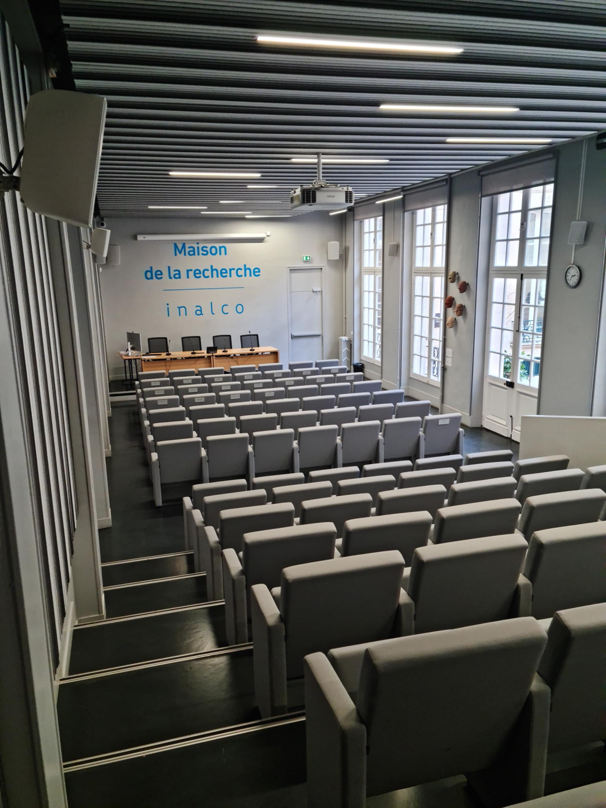Salle de conférence avec rangées de chaises, table, projecteur, écran et horloge.