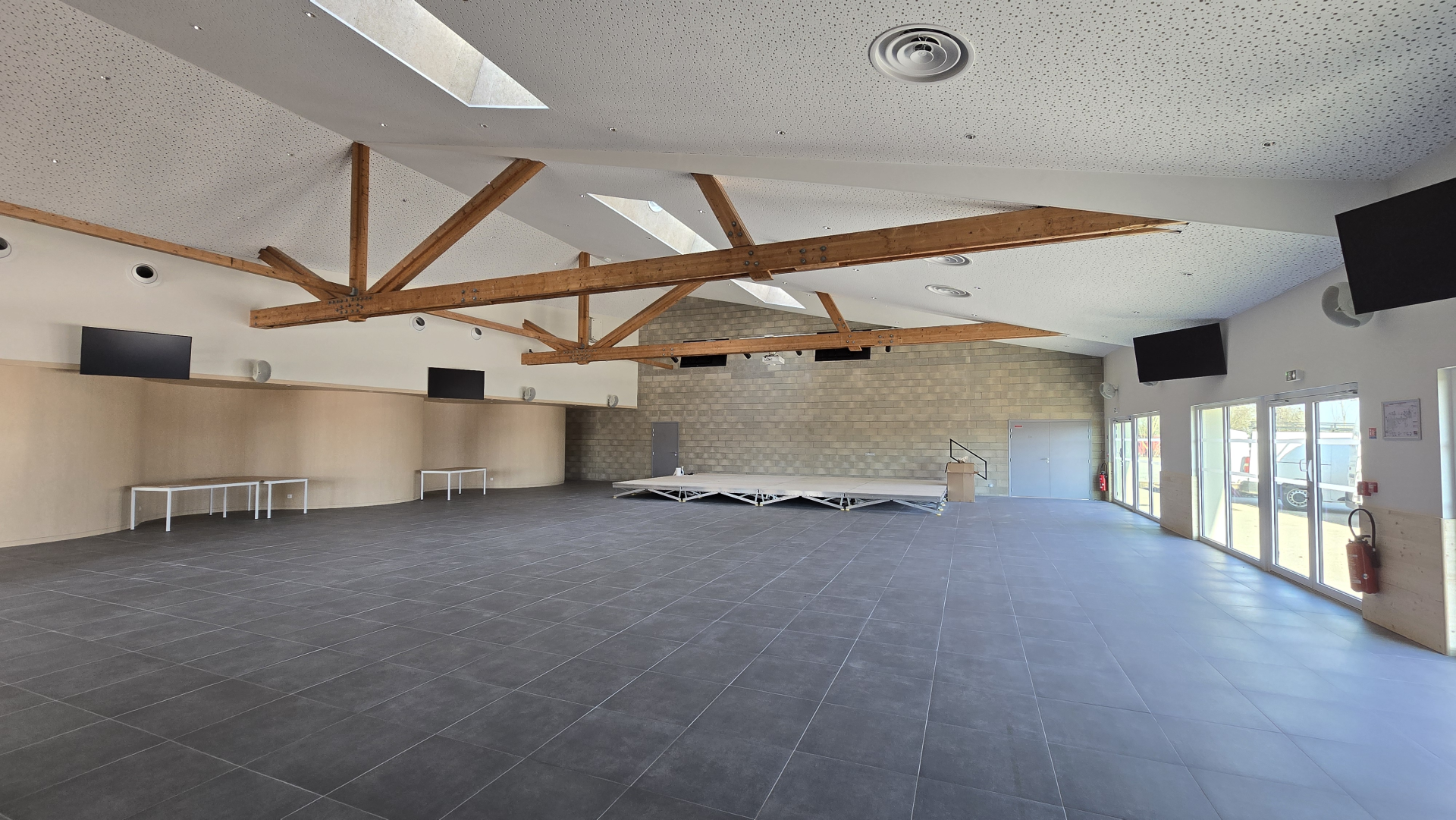 Salle avec poutres en bois, écrans, tables pliables et rangées contre le mur.