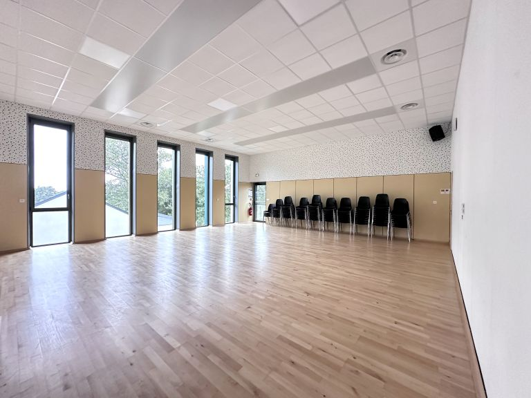 Salle spacieuse avec parquet, chaises alignées, grandes fenêtres et murs en bois clair.