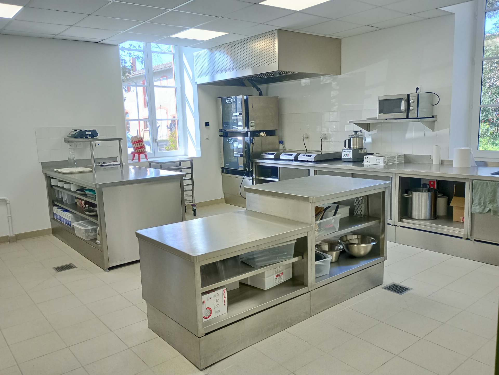 Cuisine professionnelle avec plans de travail, étagères, évier, réfrigérateur, micro-ondes, hotte et divers équipements.