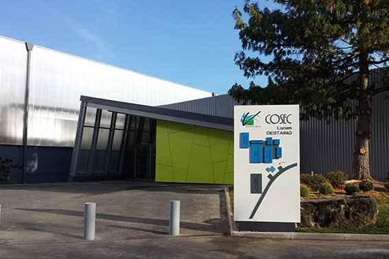 Bâtiment avec entrée, panneau directionnel, poteaux de parking, et arbre.