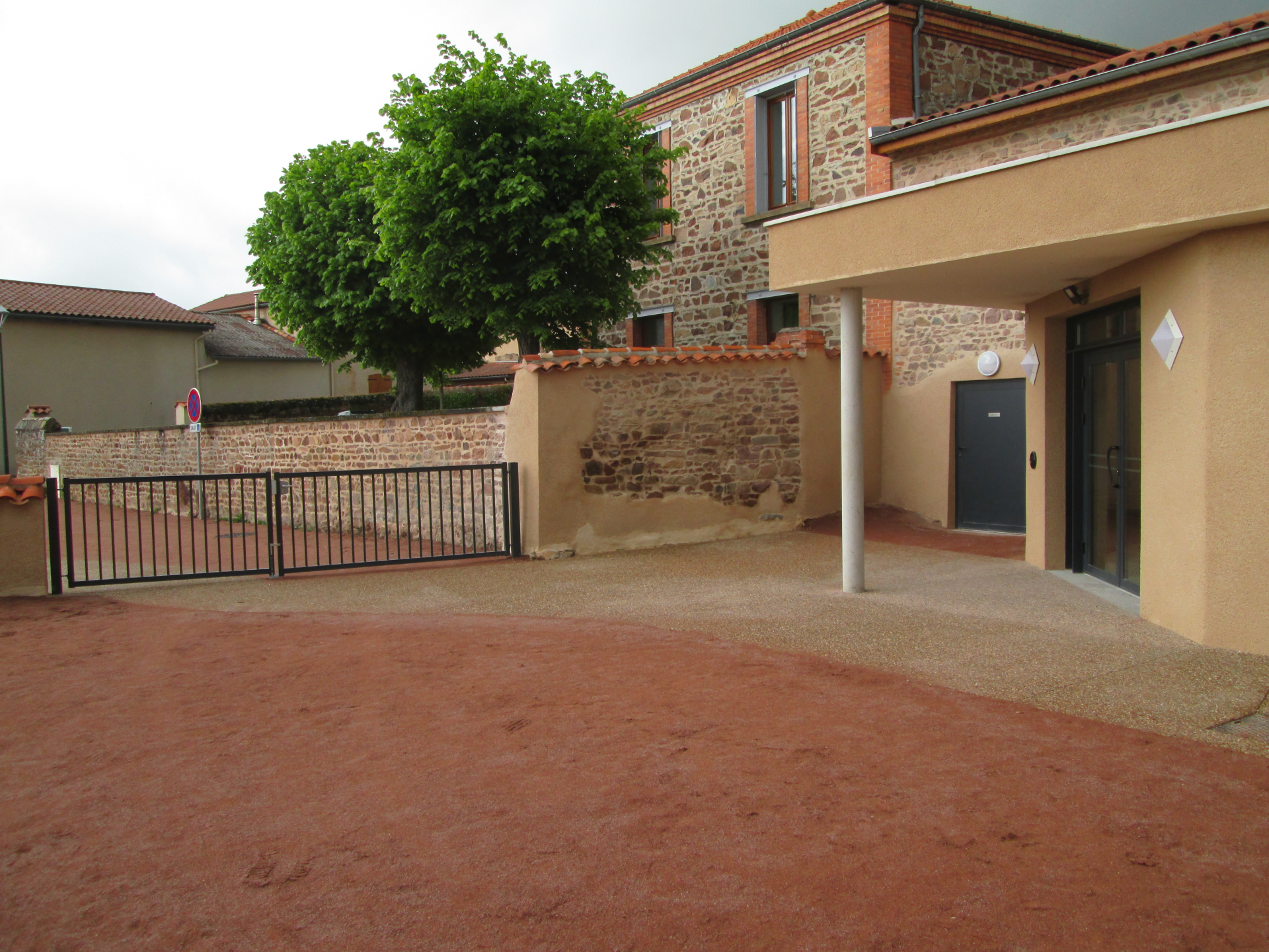 Cour avec sol en terre battue, bâtiment en pierre et briques, portail métallique, arbre.