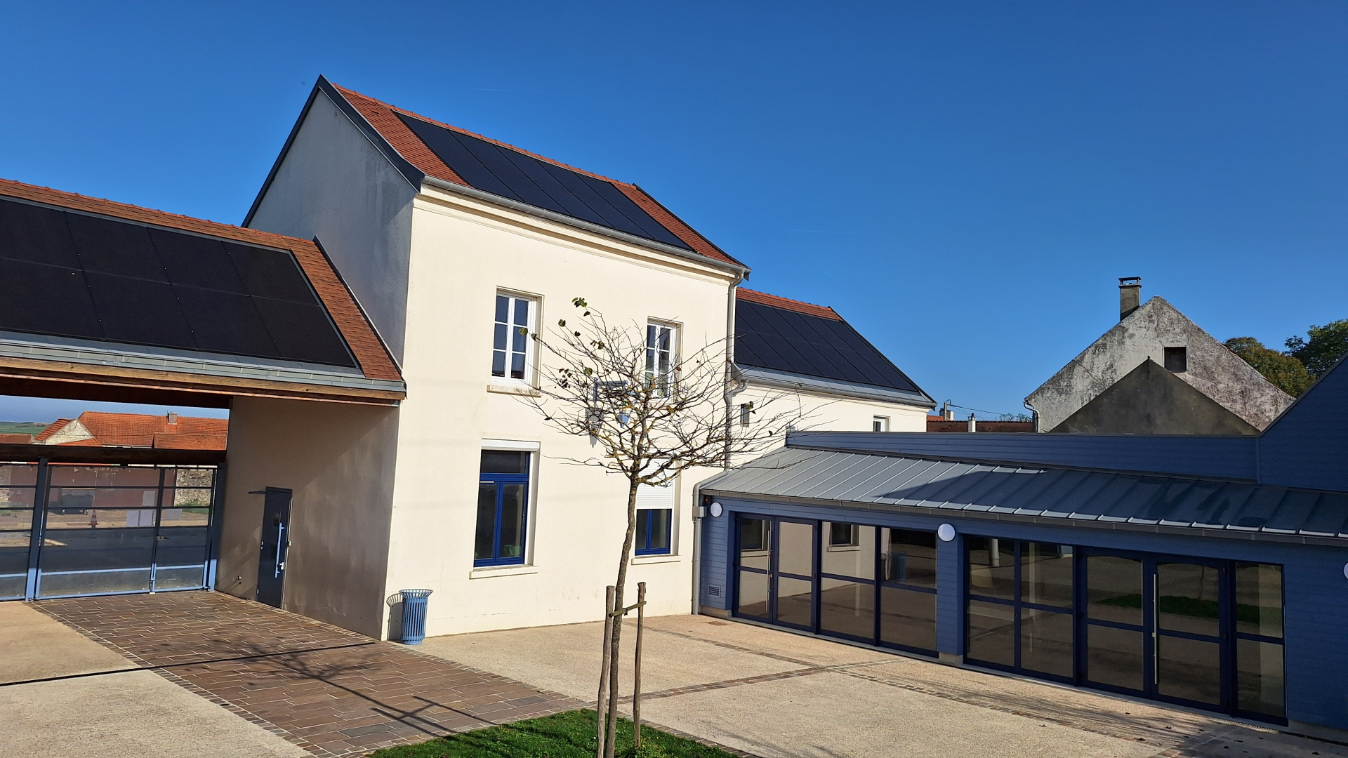 Bâtiment avec toits en pente, panneaux solaires, fenêtres, porte, arbre, poubelle, et extension moderne avec grandes vitres.