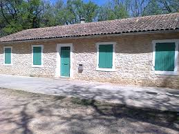 Bâtiment de pierre avec porte verte centrale, trois fenêtres vertes, allée cimentée et arbre à l'avant.