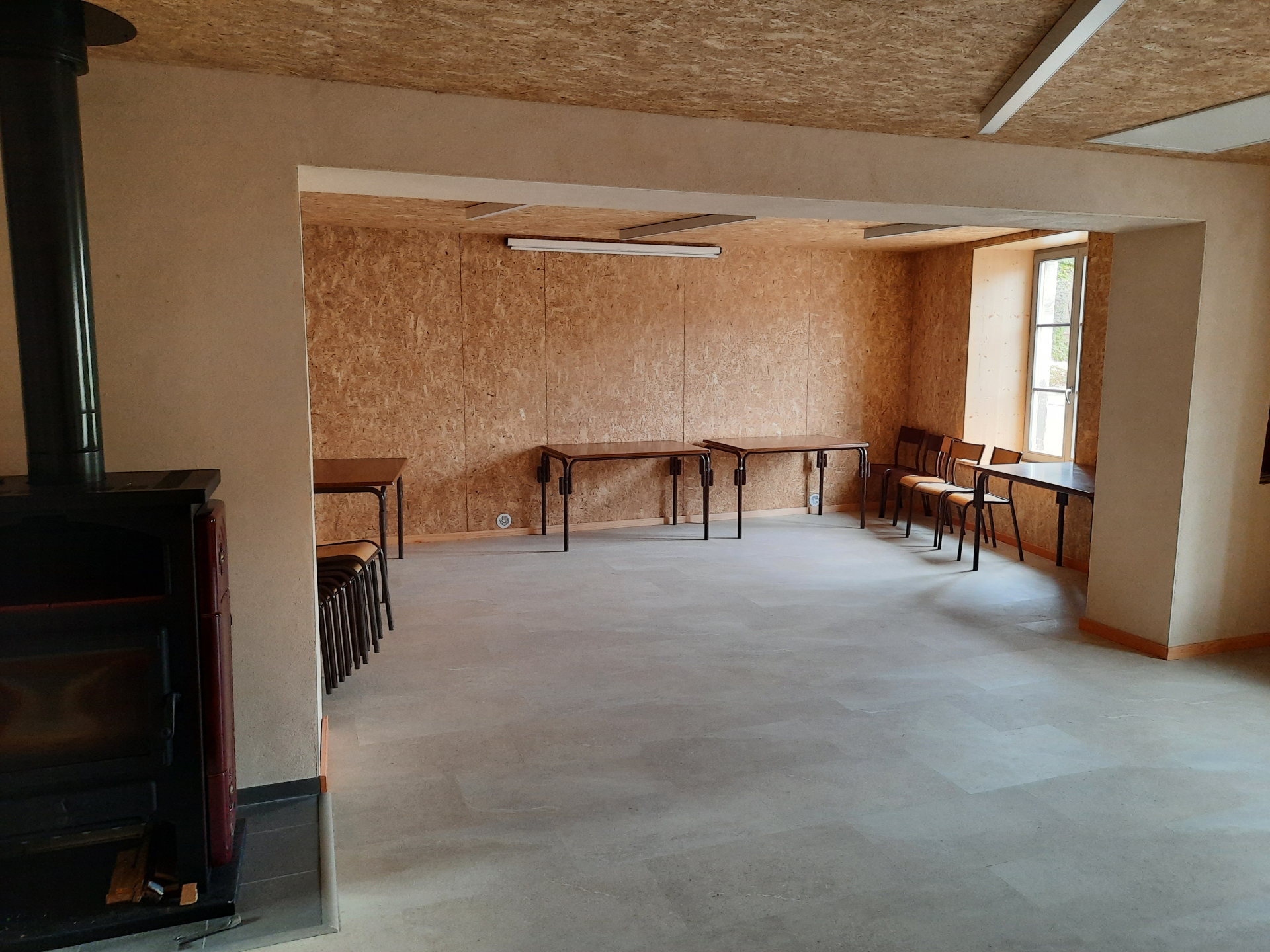 Pièce avec tables, chaises, poêle à bois et fenêtre.