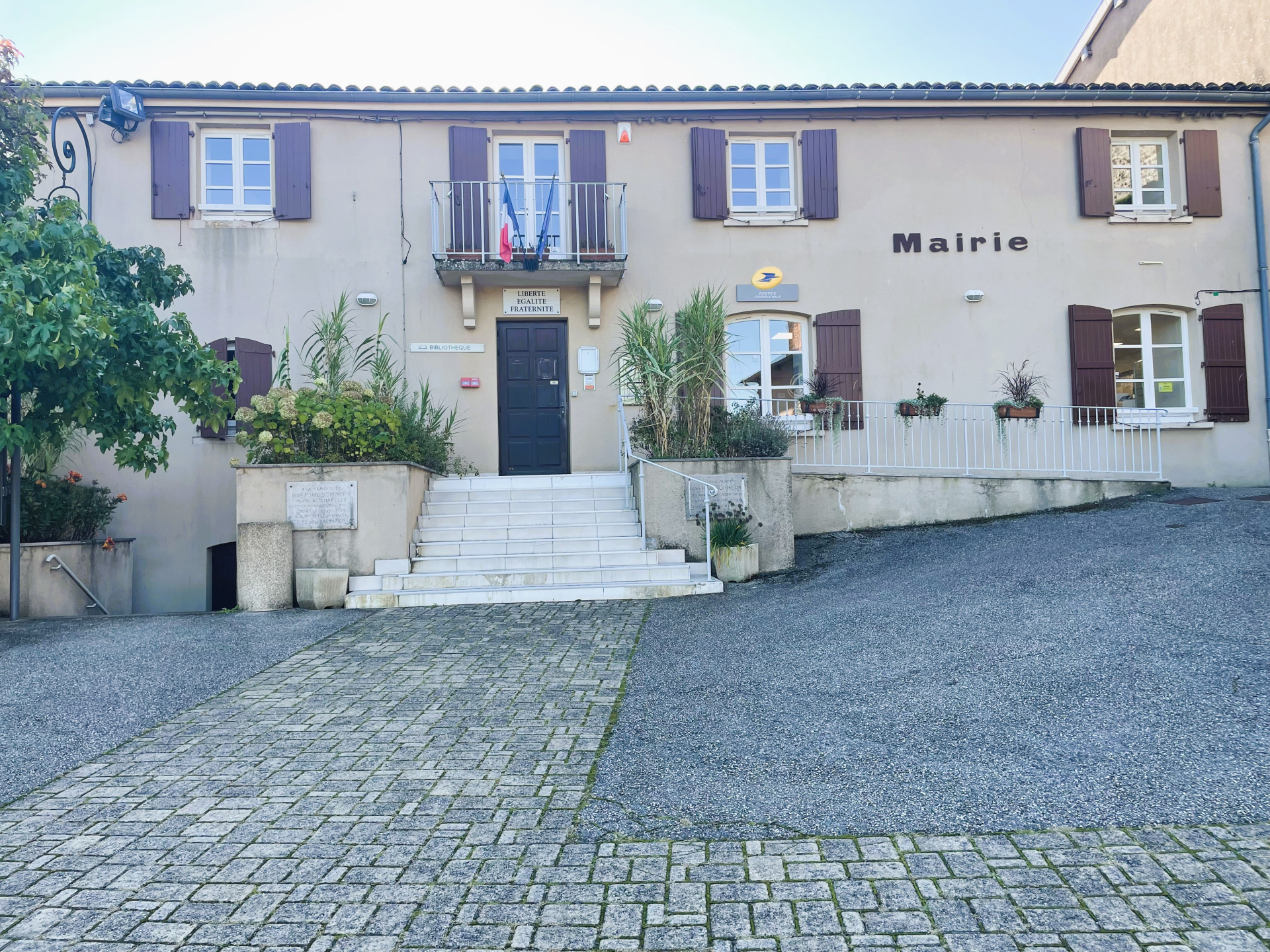 Mairie avec façade, volets, pots de fleurs, escalier et trottoir pavé.