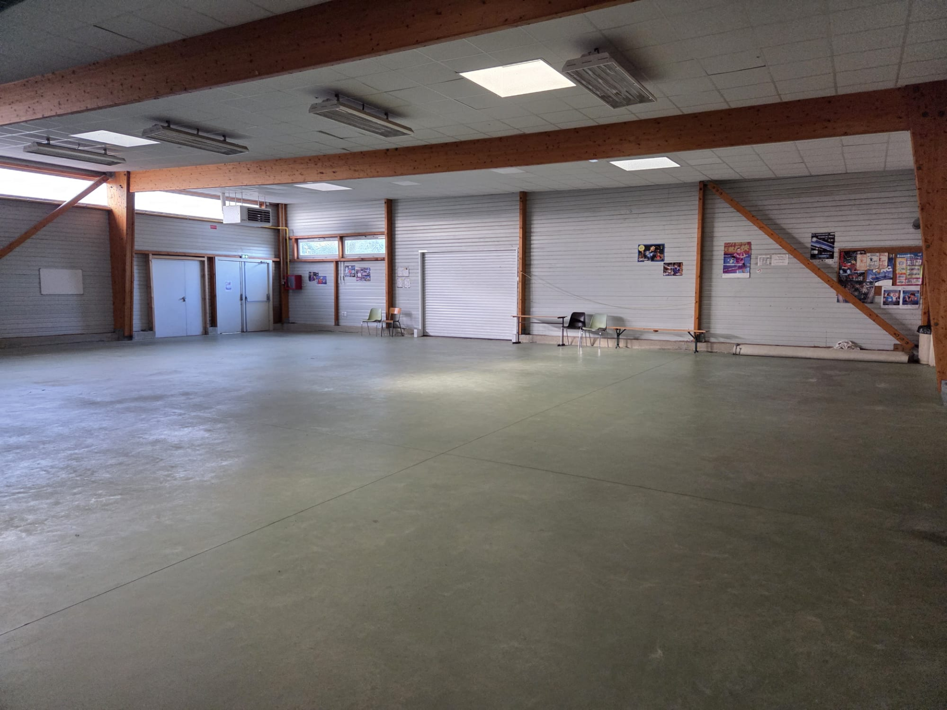 Salle vide avec sol en béton, tables, chaises, portes et affiches murales.