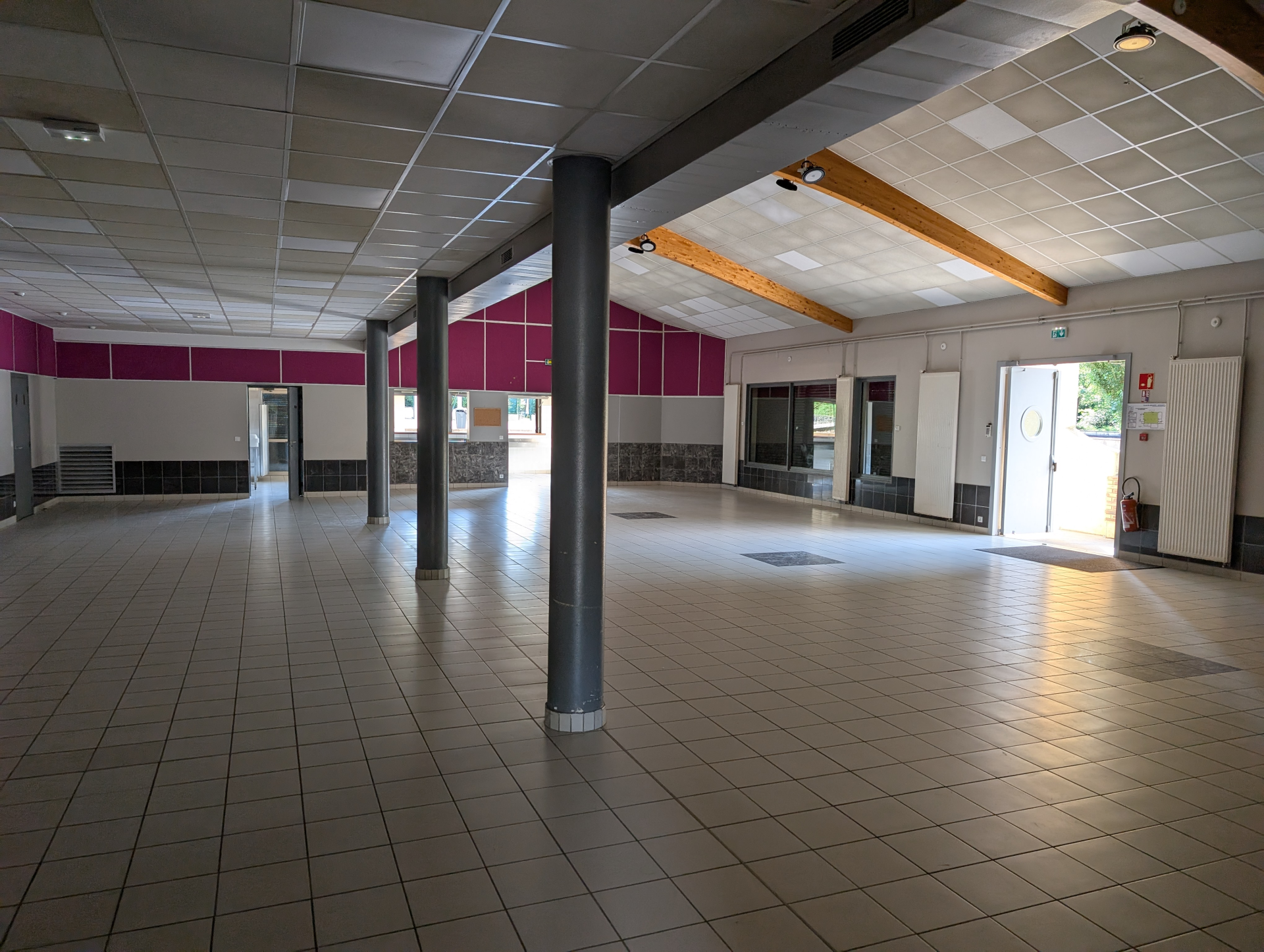 Grand espace vide avec colonnes, portes, fenêtres et sorties de secours. Sol carrelé, murs rose et plafond avec dalles.