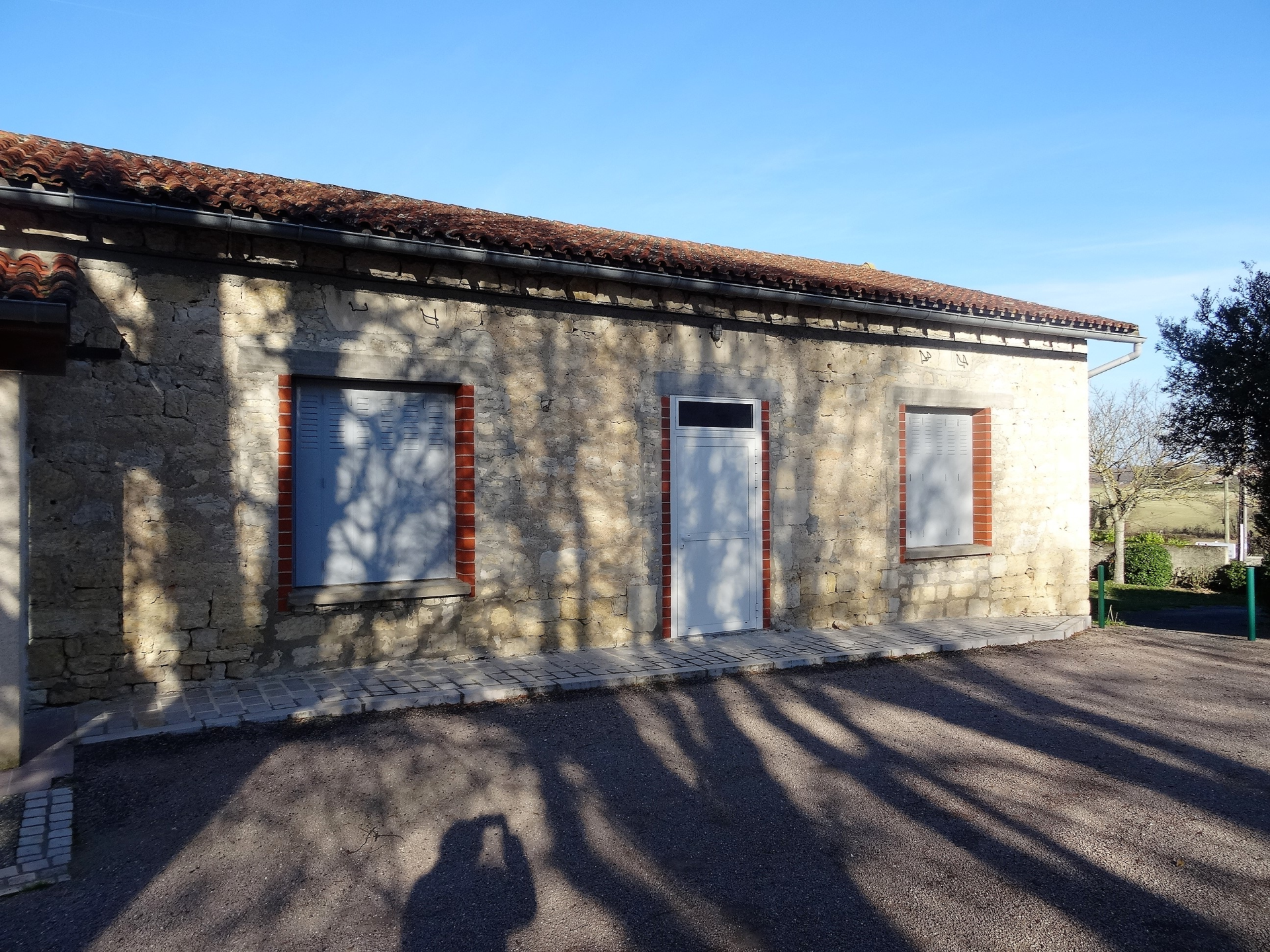 Bâtiment en pierre avec porte blanche et deux fenêtres aux volets rouges.
