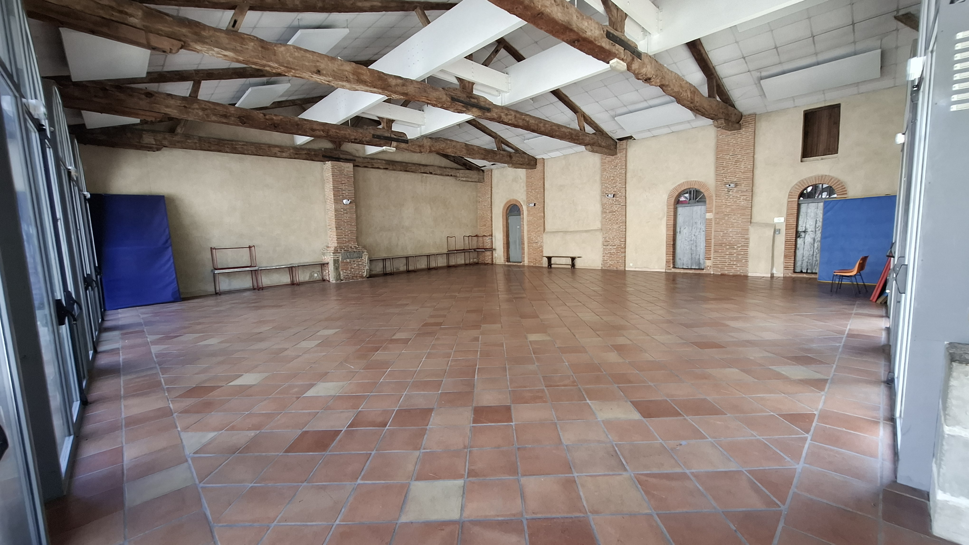 Salle spacieuse avec sol carrelé, poutres apparentes, bancs, portes et fenêtres.