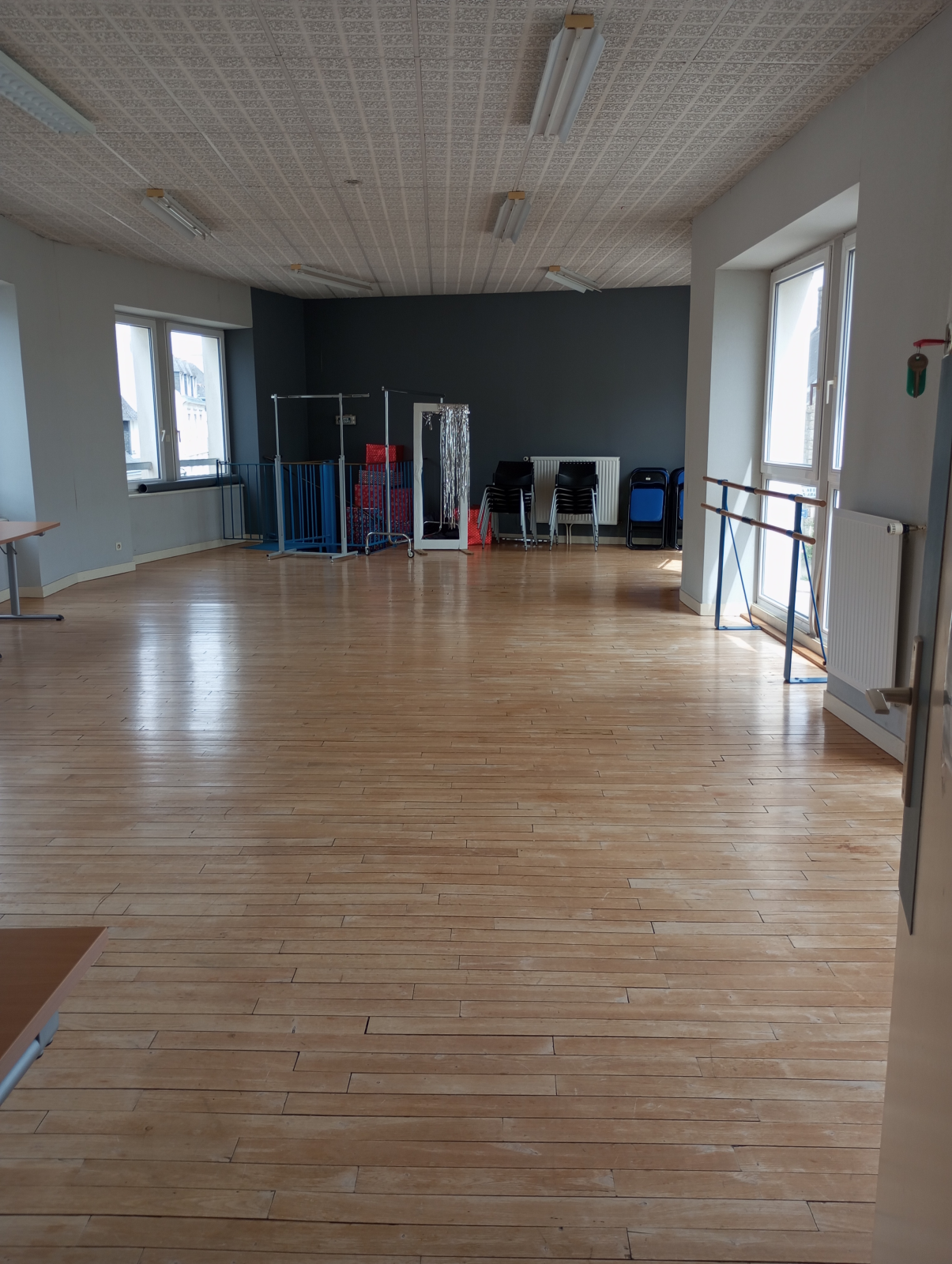 Salle vide avec parois vitrées, plancher en bois, barre d'appui, bancs, et matériel de sport.