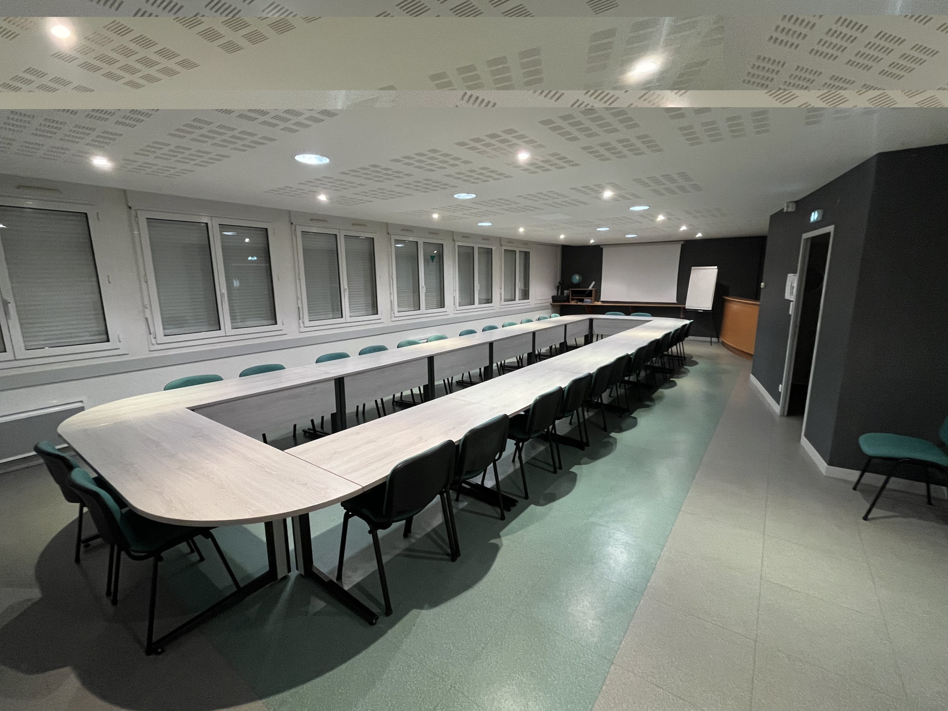 Salle de réunion avec table en U, chaises, écran, vidéo projecteur et ordinateur.