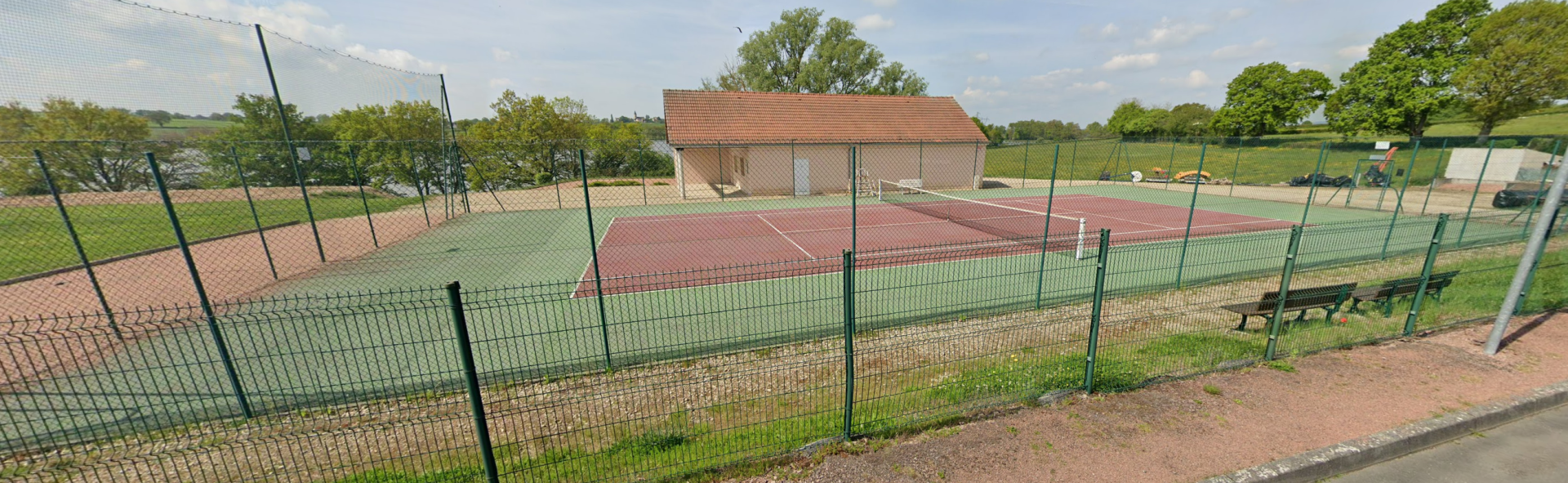 Terrain de tennis avec clôture, filet et banc. Bâtiment adjacent.