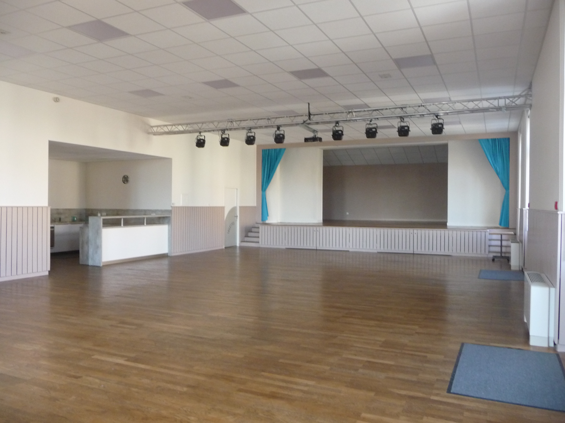 Salle spacieuse avec projecteurs, scène, comptoir et plancher en bois.