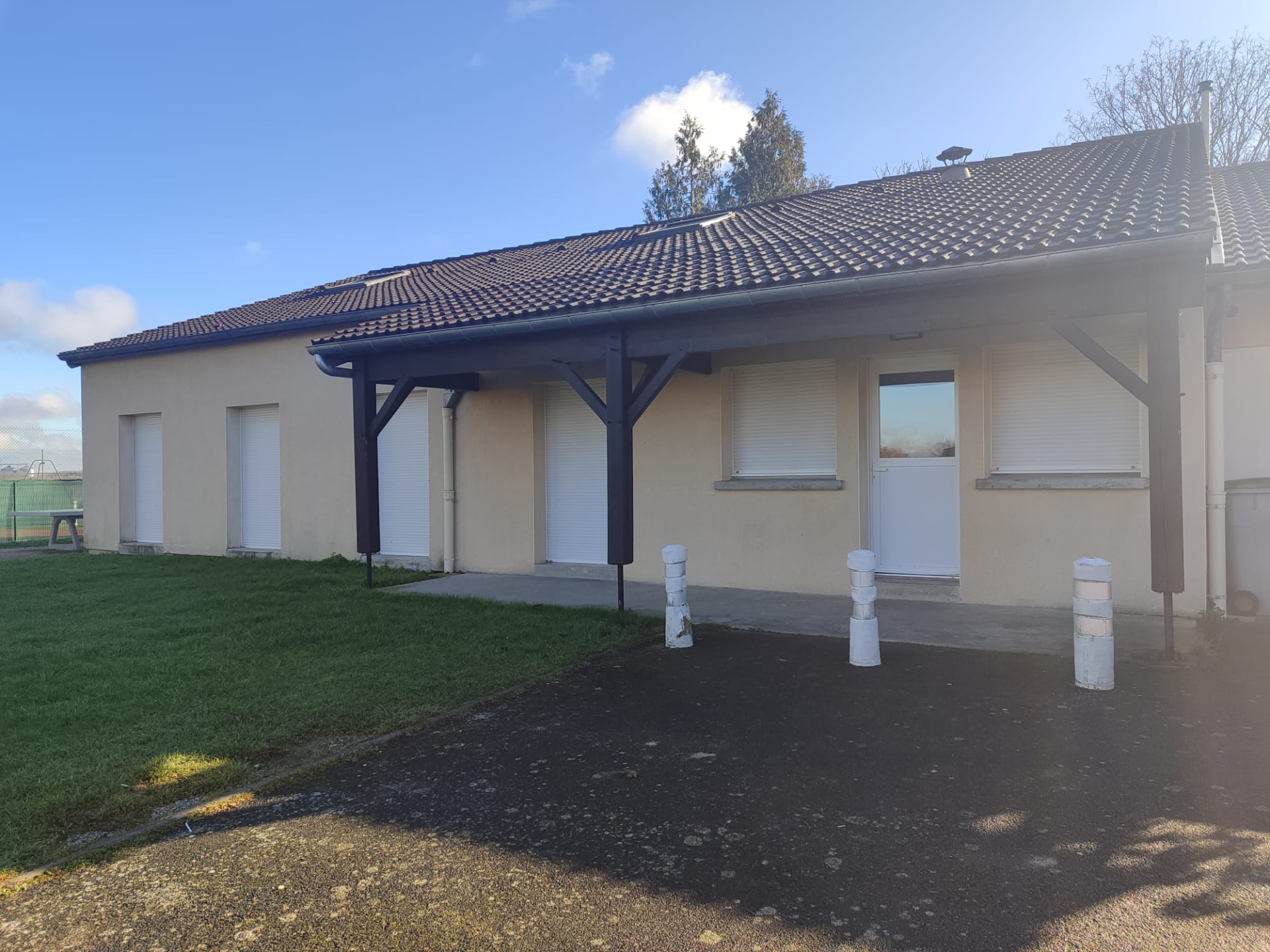 Bâtiment à toit en tuiles, murs crème, portes et fenêtres blanches. Poteaux blancs devant. Pelouse à gauche.