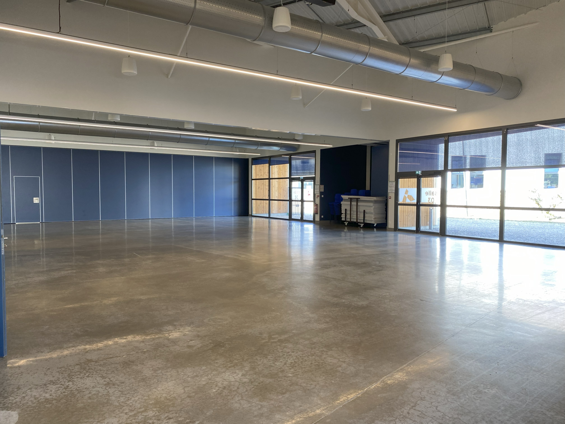 Grande salle vide avec sol en béton, grandes fenêtres, murs bleus et portiques suspendus au plafond.