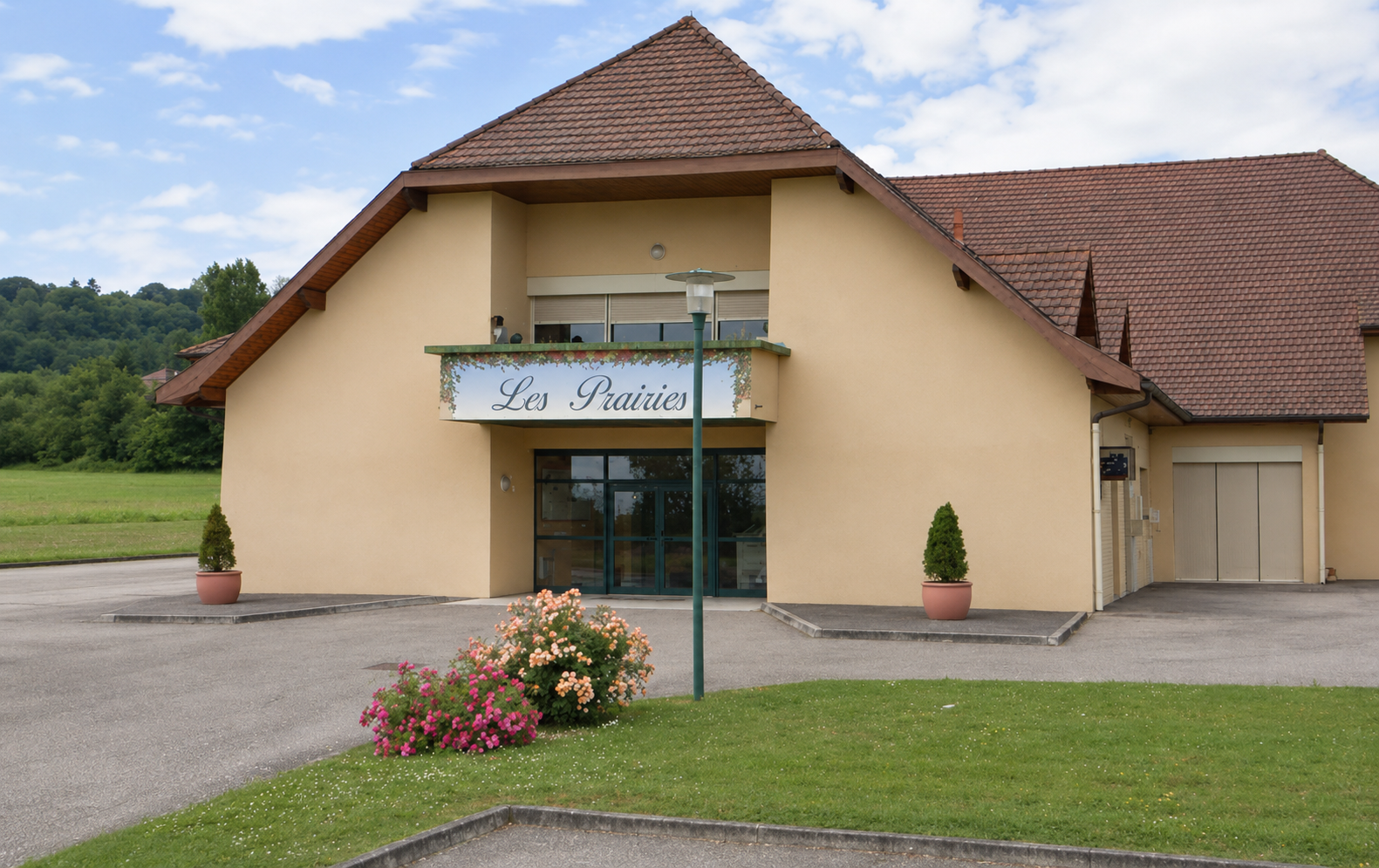 Bâtiment avec enseigne "Les Prairies", entrée vitrée, deux pots de fleurs, pelouse et parking.