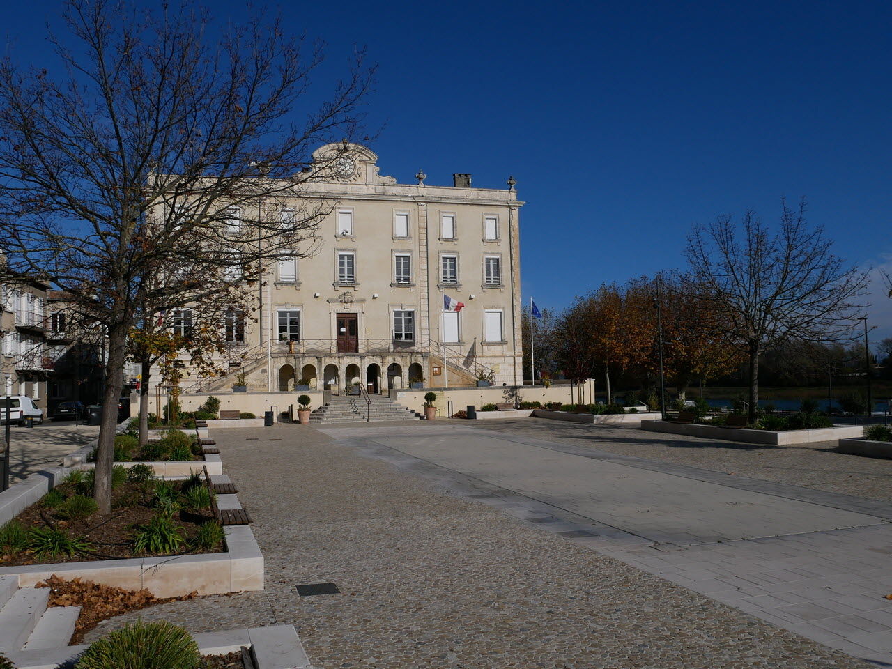 Place devant un bâtiment avec arbres, végétation et allée pavée.