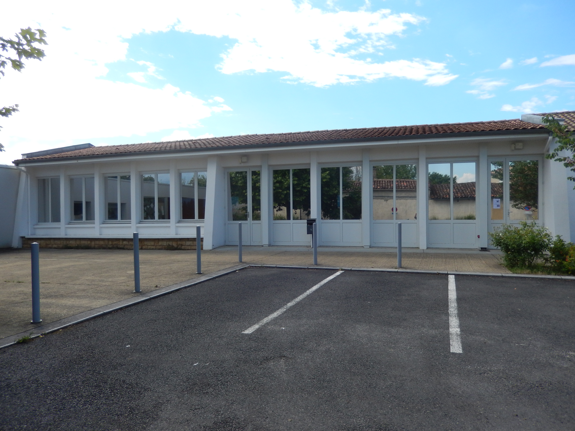 Bâtiment avec grandes fenêtres, toit en tuiles, parking avec places marquées.