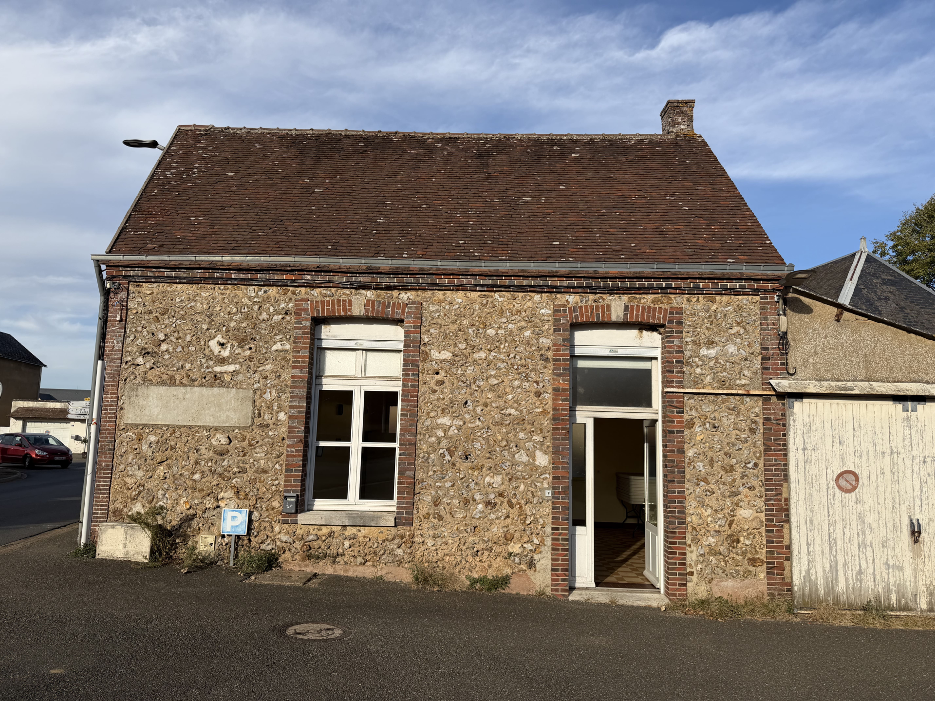 Bâtiment en pierre avec toit en tuiles, porte blanche ouverte, fenêtre à droite, porte de garage blanche à gauche.