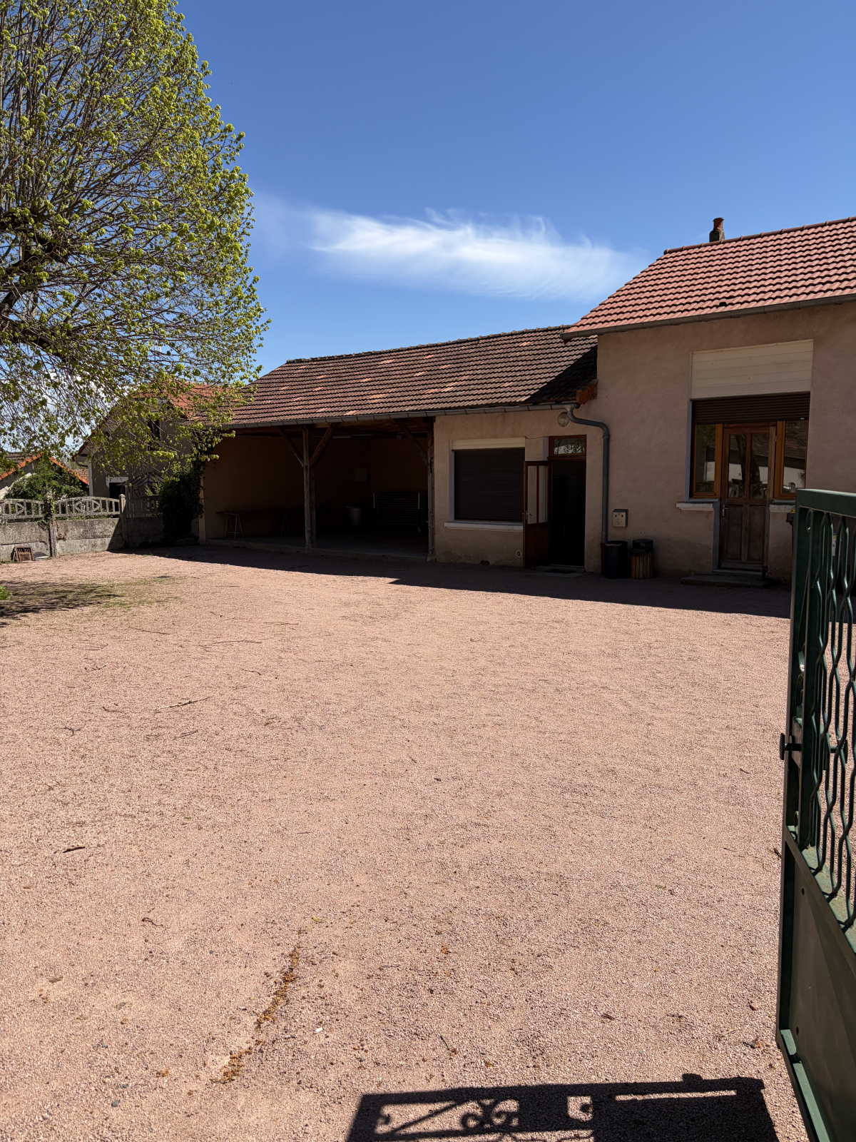 Maison avec cour en gravier, arbre, portail et barbecue.