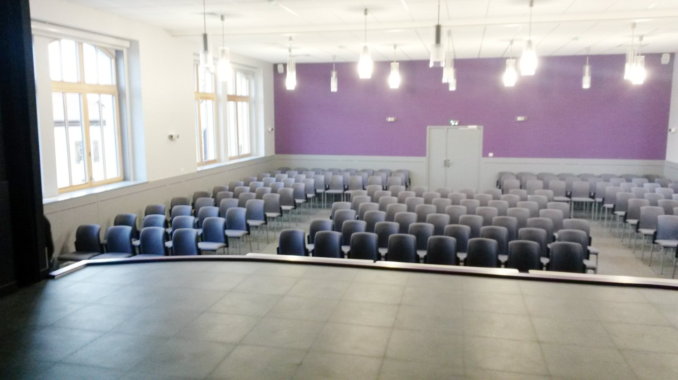 Salle avec rangées de chaises, scène, fenêtres, portes et éclairage suspendu.