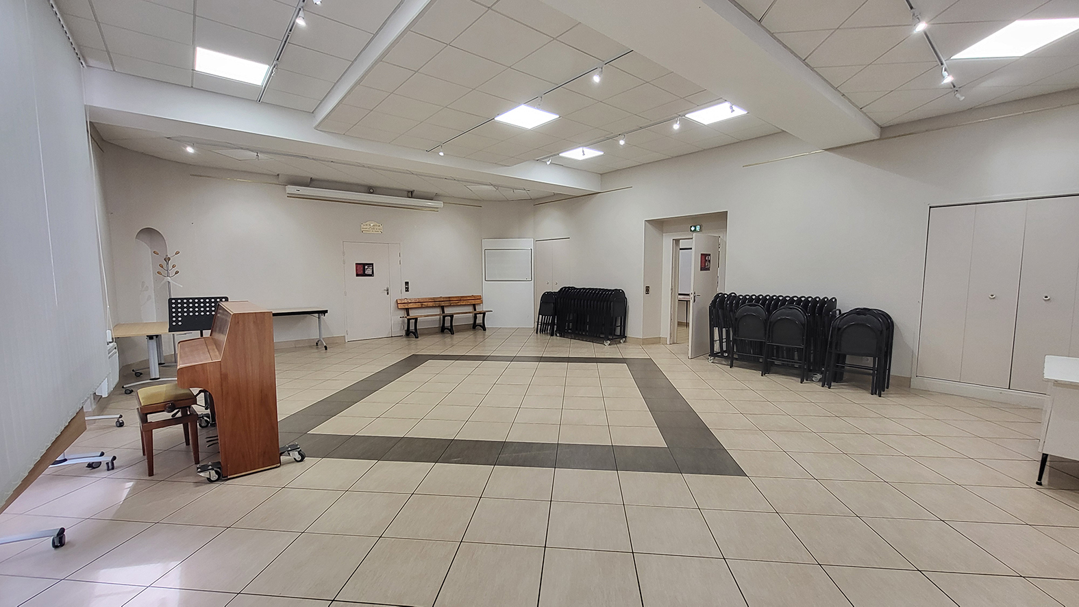 Salle avec piano, pupitre, tables, chaises, tableau blanc et porte.
