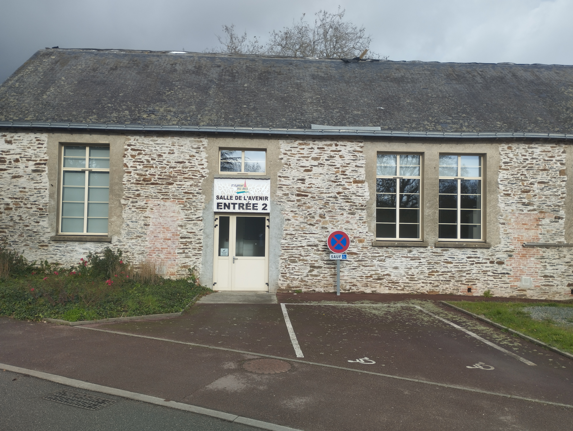 Bâtiment en pierre avec entrée marquée "Salle de l'Entente Entrée 2", parking avec places handicapées, panneau d'interdiction de tourner à gauche.
